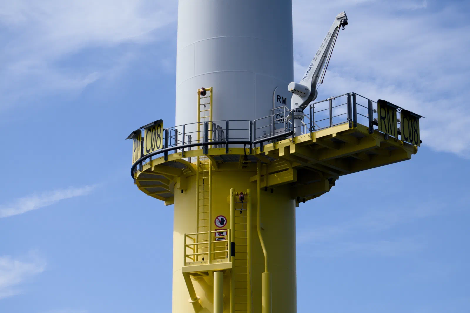 Close-up of an offshore wind turbine base with the yellow access platform, ladder and a service crane, marked RM C08, against a blue sky with thin clouds