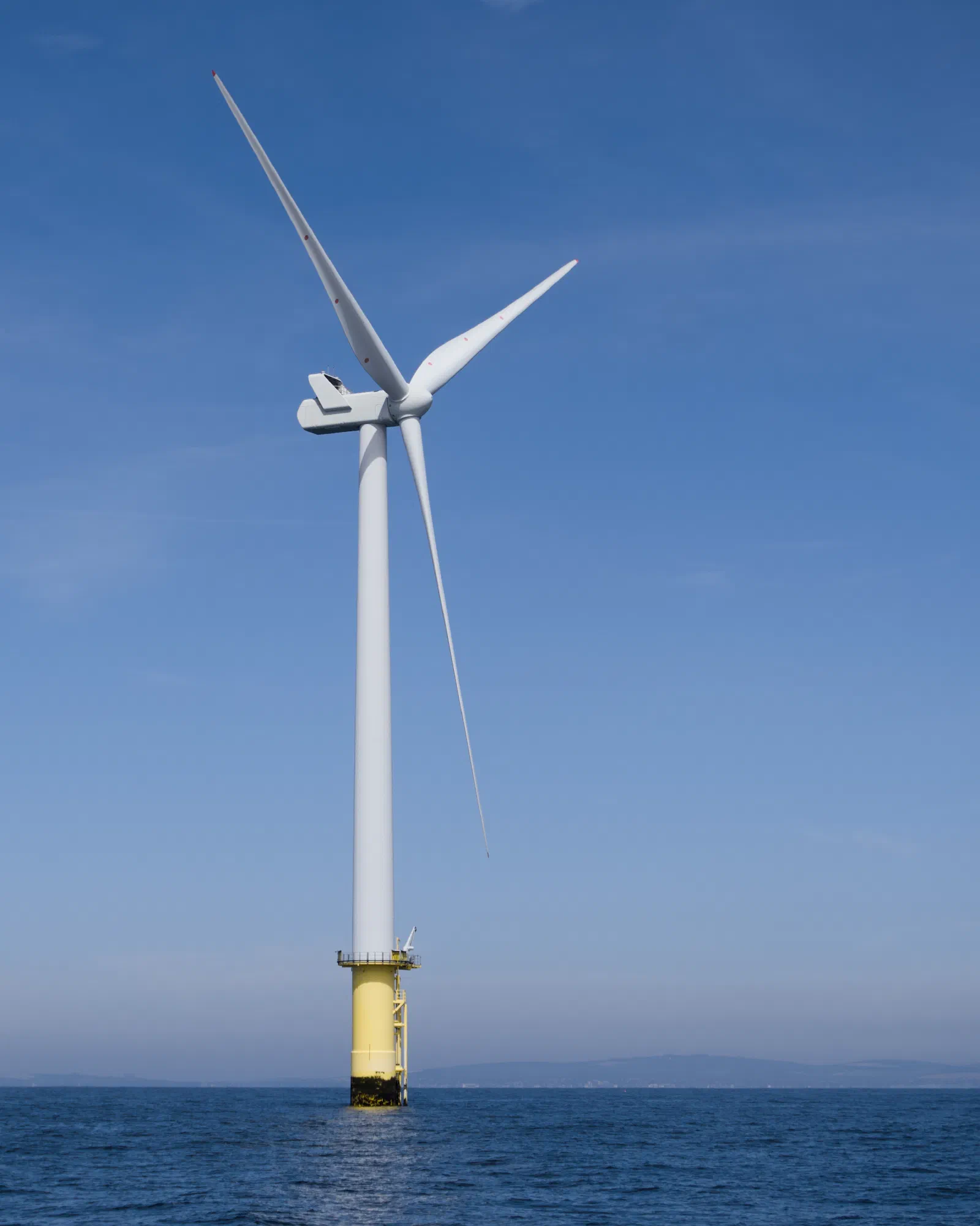 A full offshore wind turbine standing on a yellow transition piece in the sea, three white blades against a clear blue sky, the coast faintly visible on the horizon