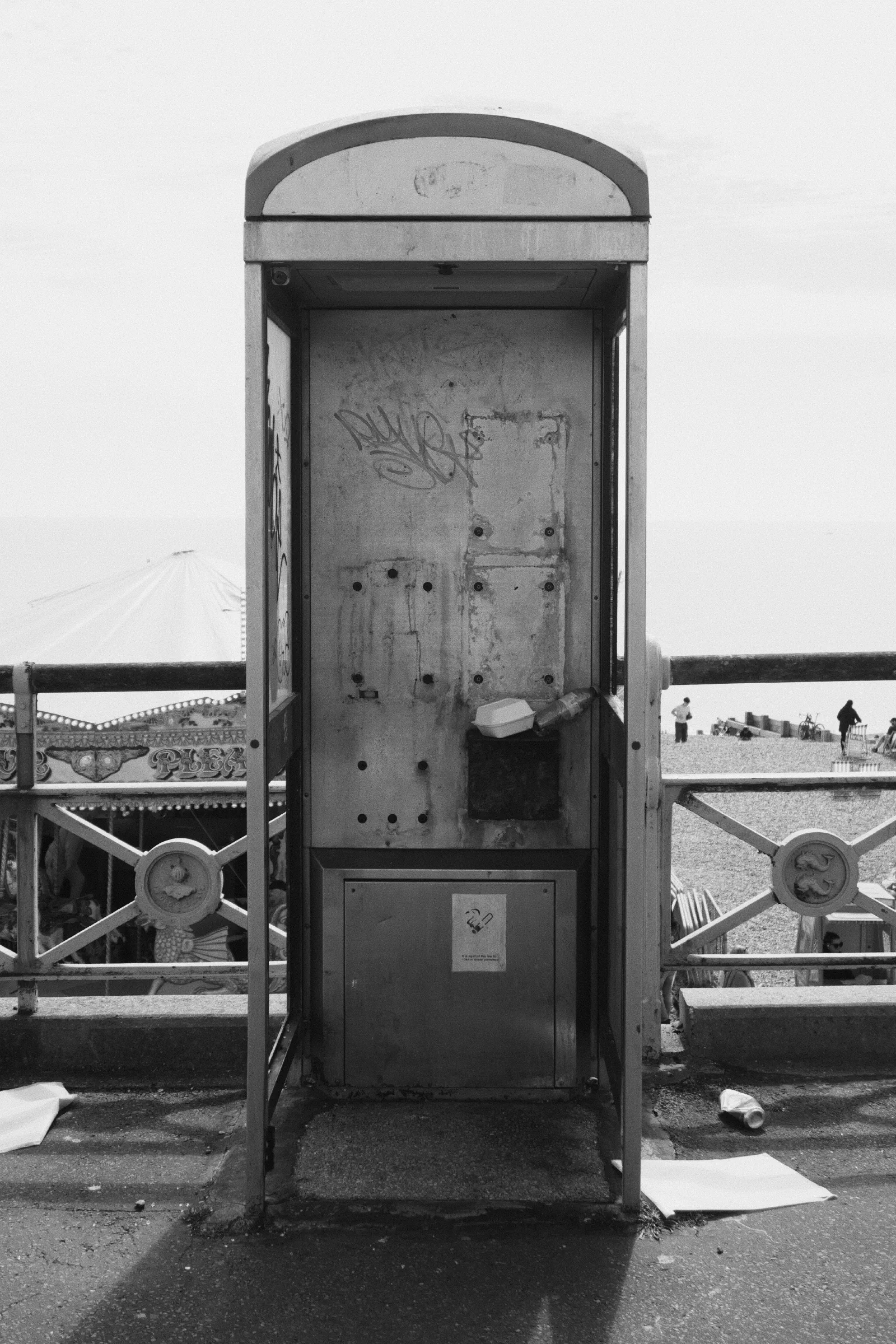 Black and white photograph of a battered, vandalised public phone booth on Brighton seafront, the receiver gone and graffiti scratched into the metal panels, beach and crowds visible behind