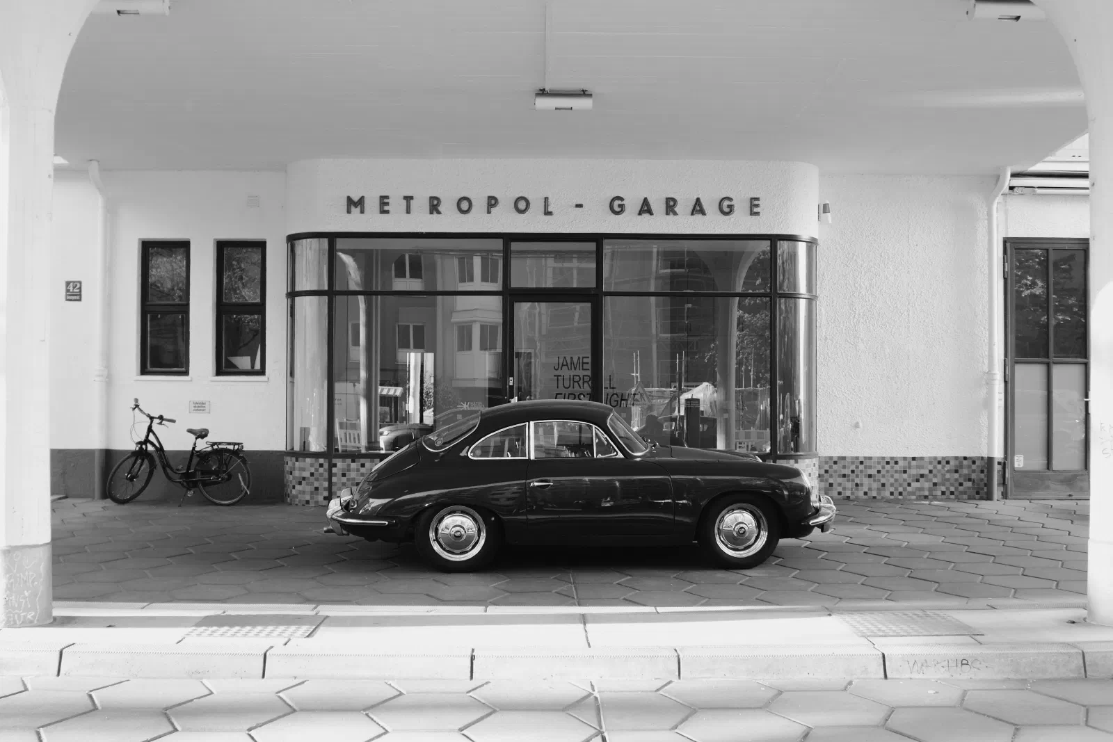 Black and white photograph of a vintage Porsche 356 parked in front of the curved glass facade of the Metropol-Garage in Munich, with a James Turrell 'First Light' exhibition poster visible through the window behind