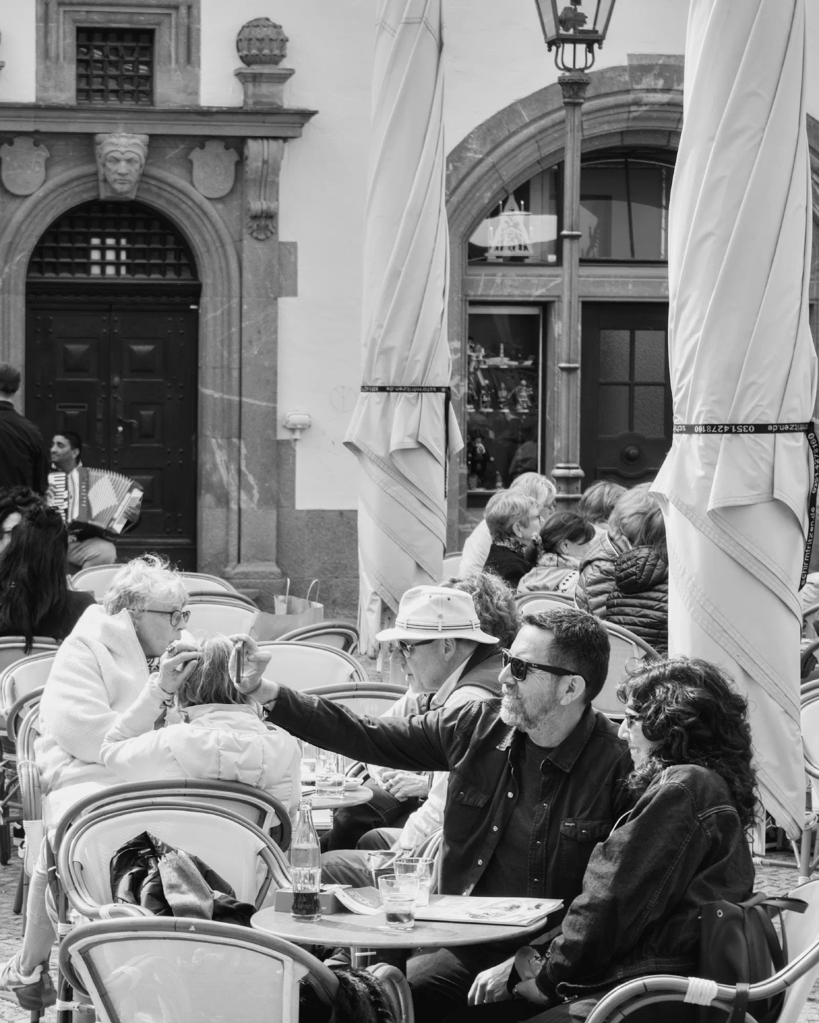 Black and white photograph of a busy outdoor cafe at Leipzig's Naschmarkt, people seated at small tables under awnings, an older woman fixing a man's hair, an accordion player and the baroque facade of the Alte Börse in the background