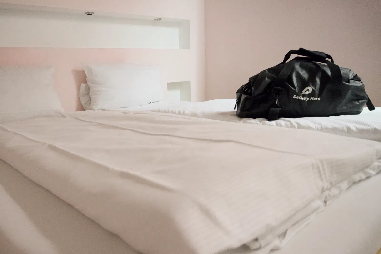 Black and white hotel room view of a freshly made bed with crisp white linen and two pillows, a black Delivery Hero duffel bag placed on the corner