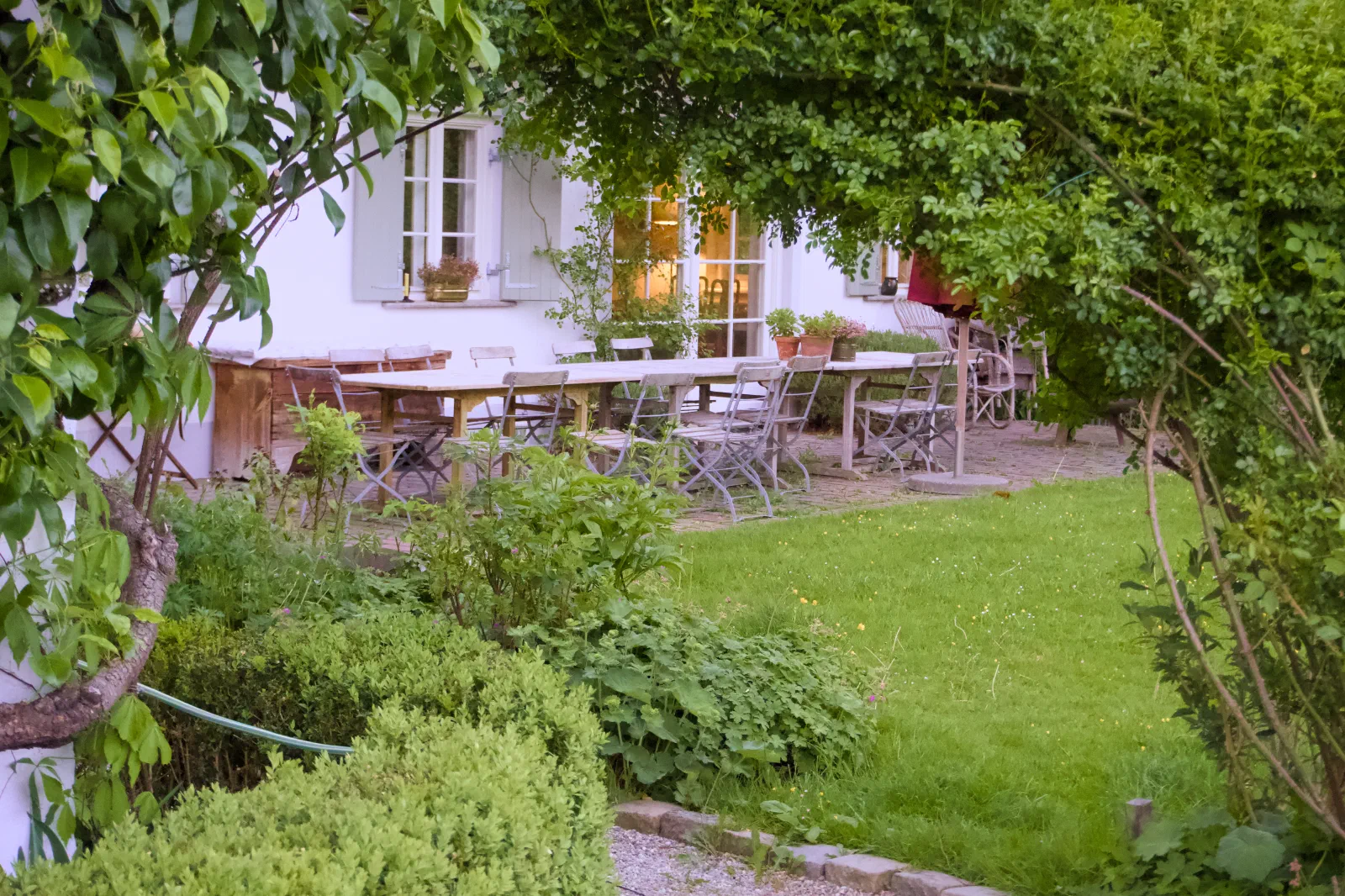 A long wooden table and folding chairs set up in a leafy garden in front of a white country house, warm light glowing from the kitchen windows in the evening