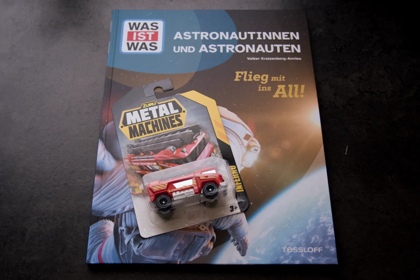 A WAS IST WAS book titled 'Astronautinnen und Astronauten - Flieg mit ins All!' lying flat with a red blister-packed Metal Machines toy car resting on top
