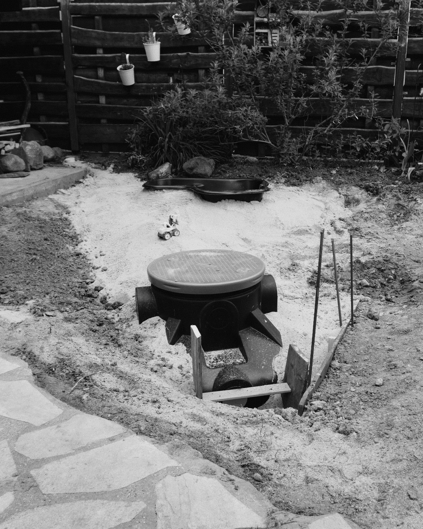 Black and white photograph of an excavated garden area with a black plastic inspection chamber sitting in a sand-filled pit, pipe stubs reaching out from the chamber, flagstones, plants and a wooden fence around it