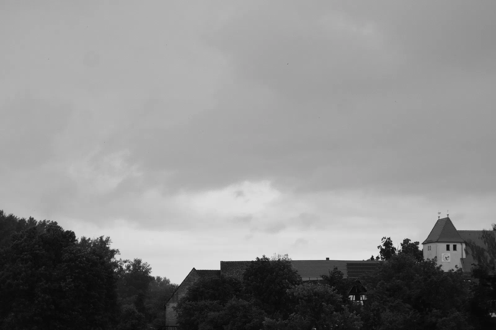 Black and white photo of a heavy overcast sky over a village, a church tower with a clock on the right above rooftops and trees