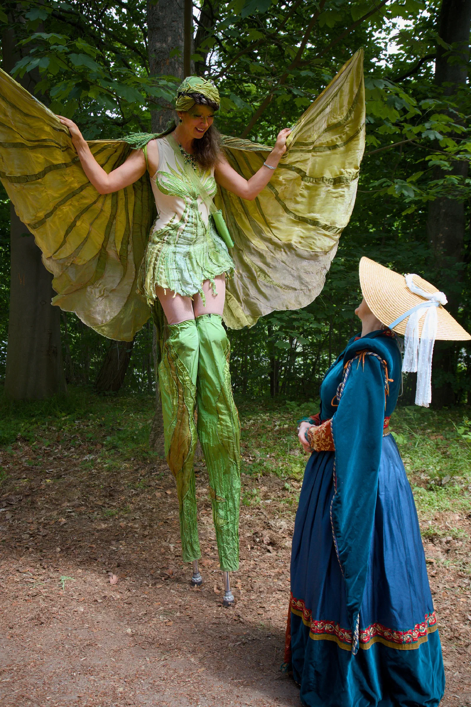 A performer on stilts in a green fairy costume talking to a woman in a straw hat and blue dress