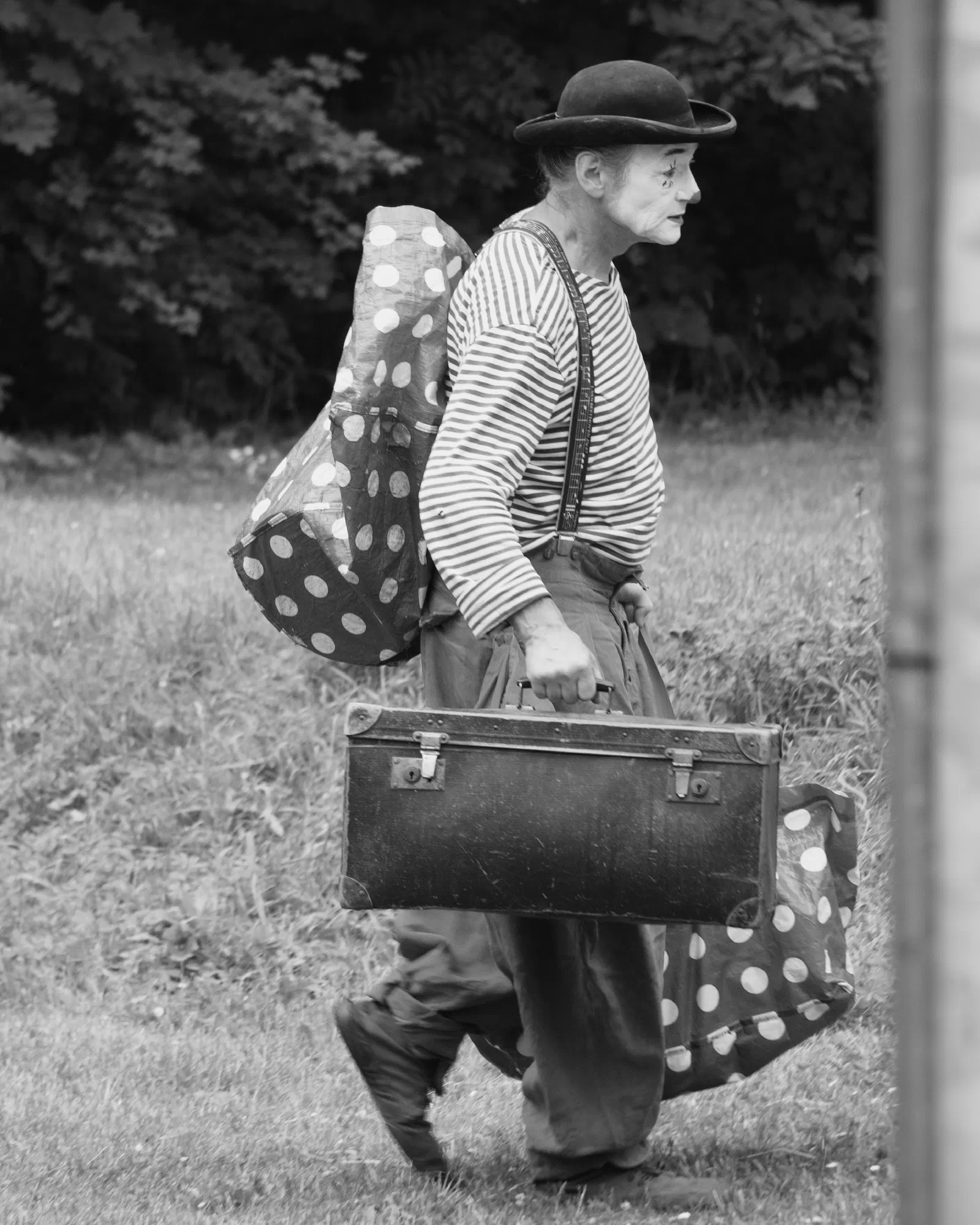 Black and white photo of a clown in a bowler hat and striped shirt walking across a meadow, carrying an old suitcase and polka-dot bundles