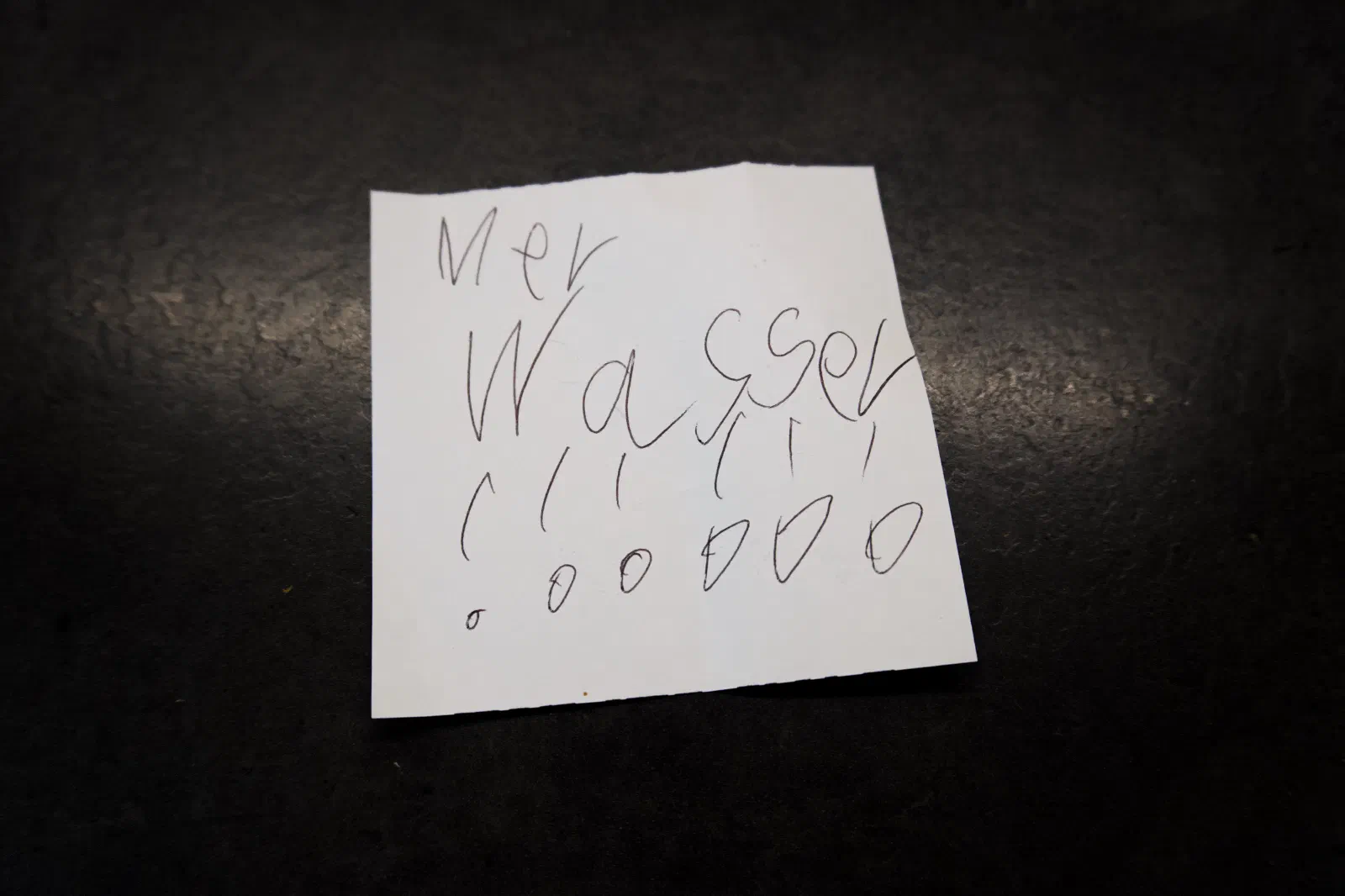 A small handwritten note on a dark surface reading 'Mer Wasser !!!' in a child's handwriting