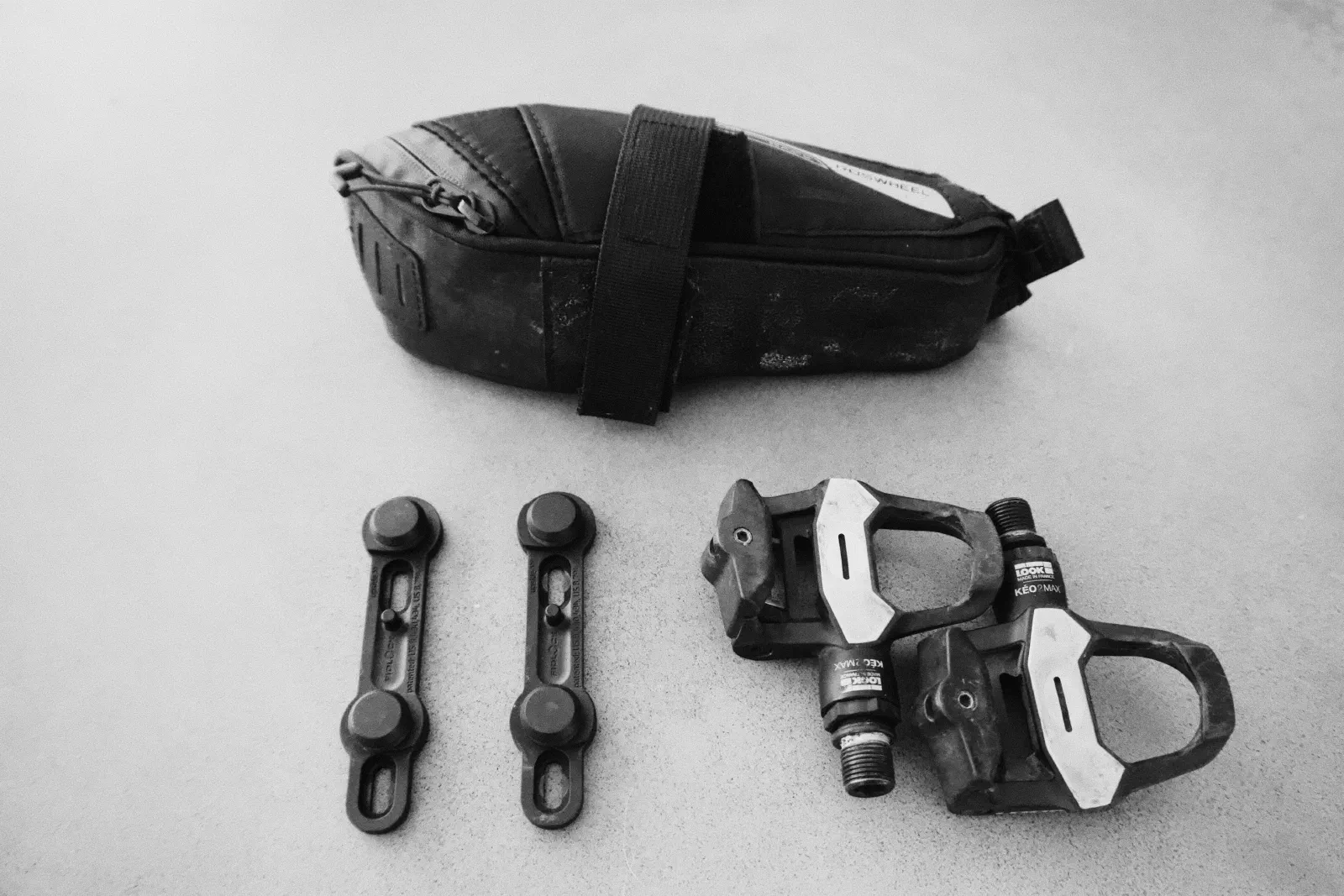 Black and white photo of a bike saddle bag, two accessory mounts, and a pair of clipless road pedals on a concrete floor