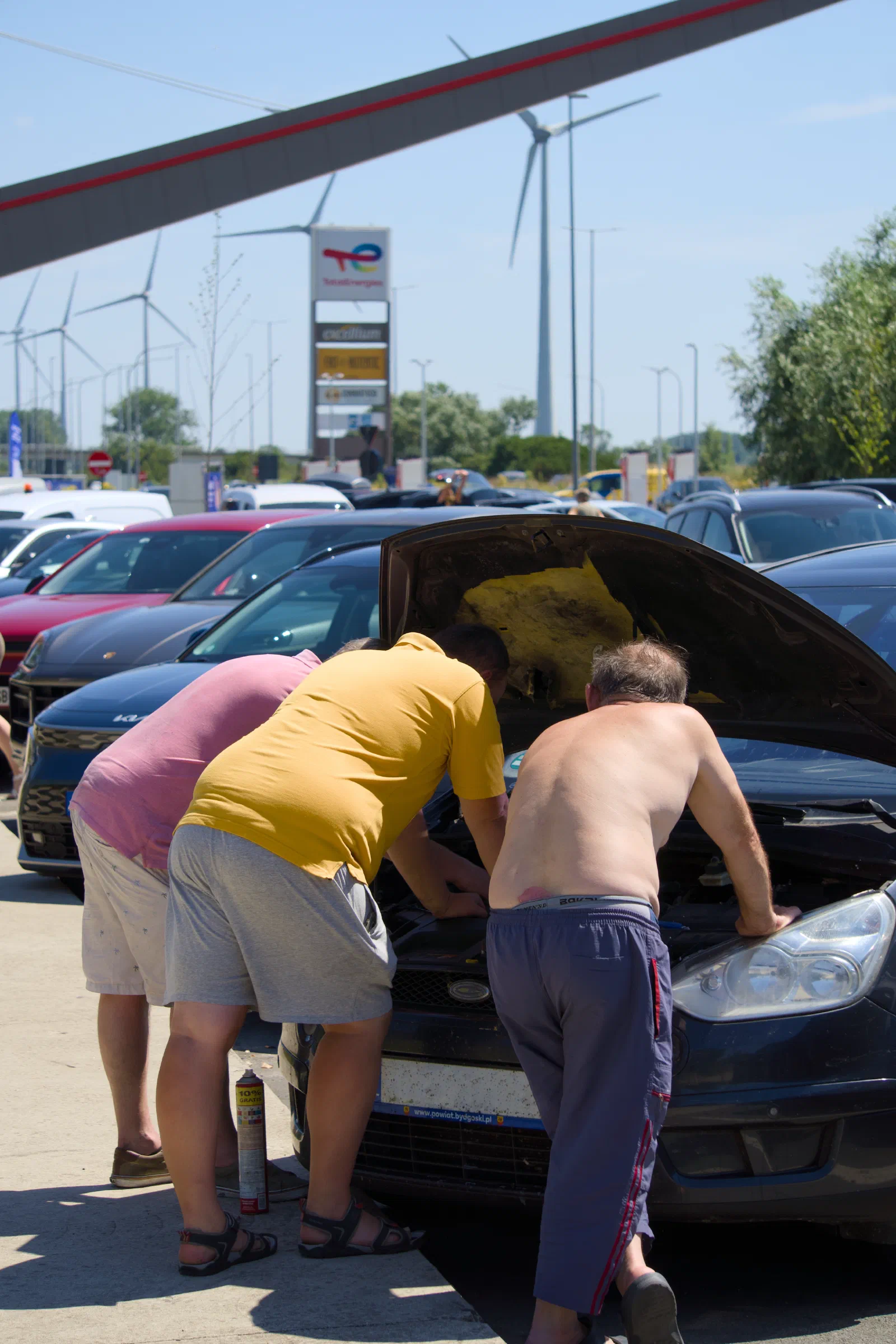 Three men under the open hood of a car at a service area, wind turbines behind
