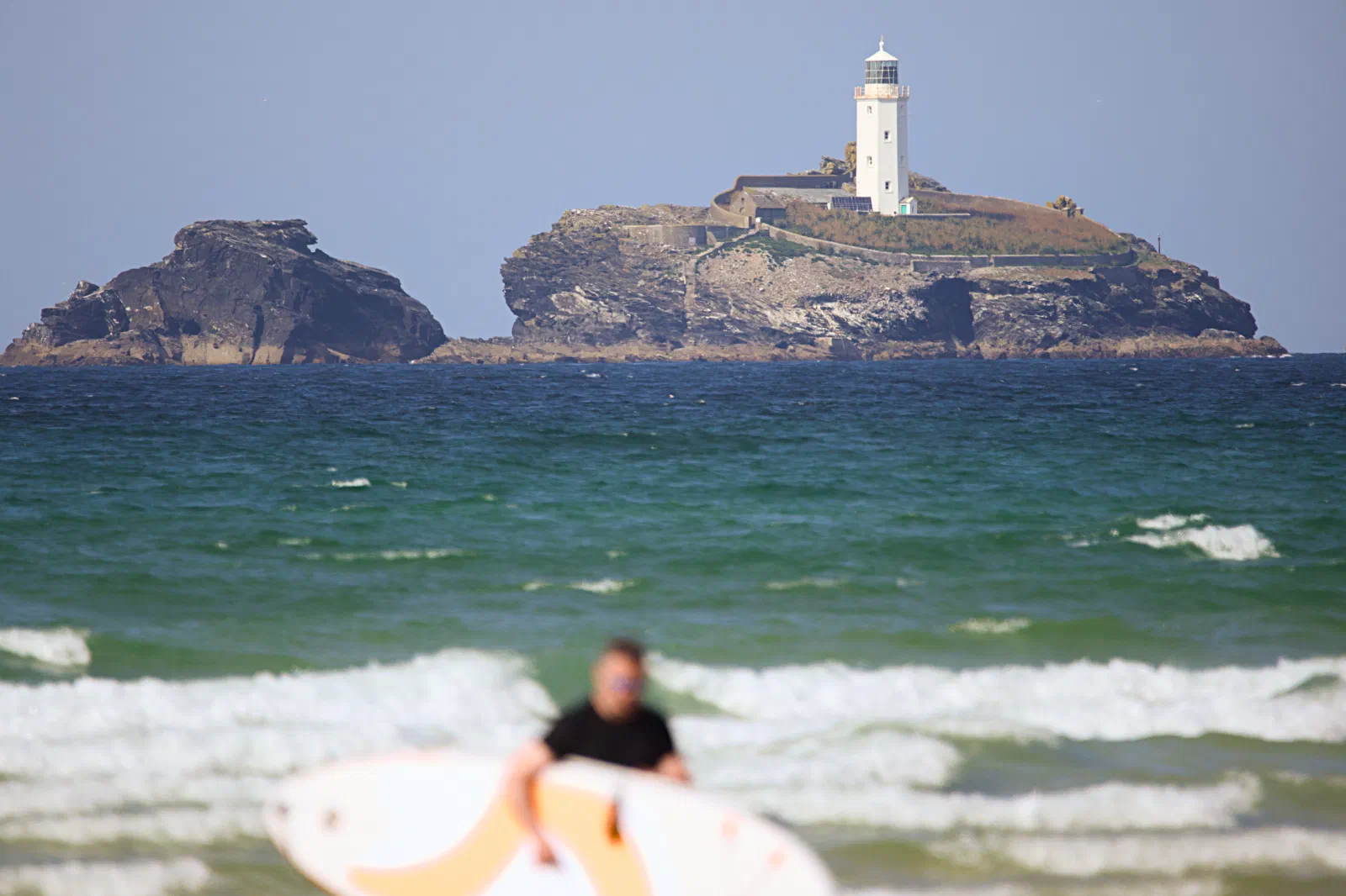 Godrevy lighthouse on its island, a surfer carrying a board in the foreground