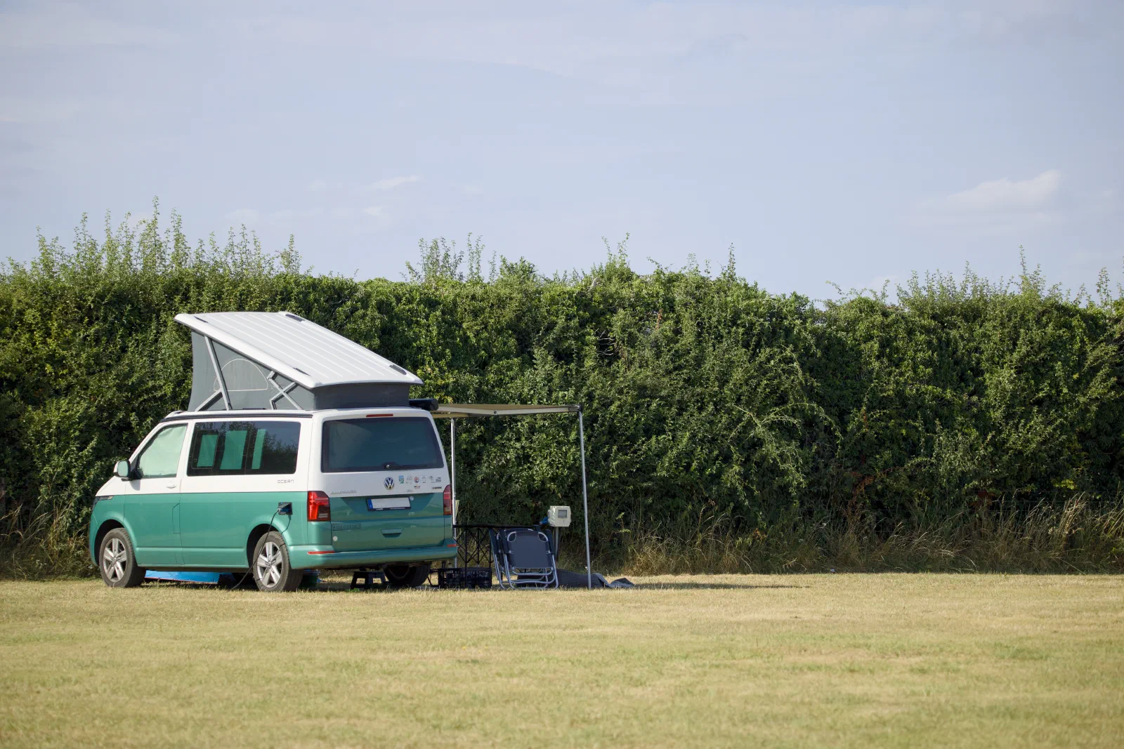 The California with the roof up and awning out on the pitch in front of a high hedge