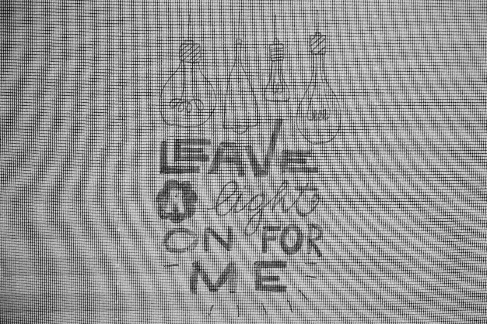 Black and white photo of a hand-drawn window message reading Leave a light on for me, with four light bulbs above it