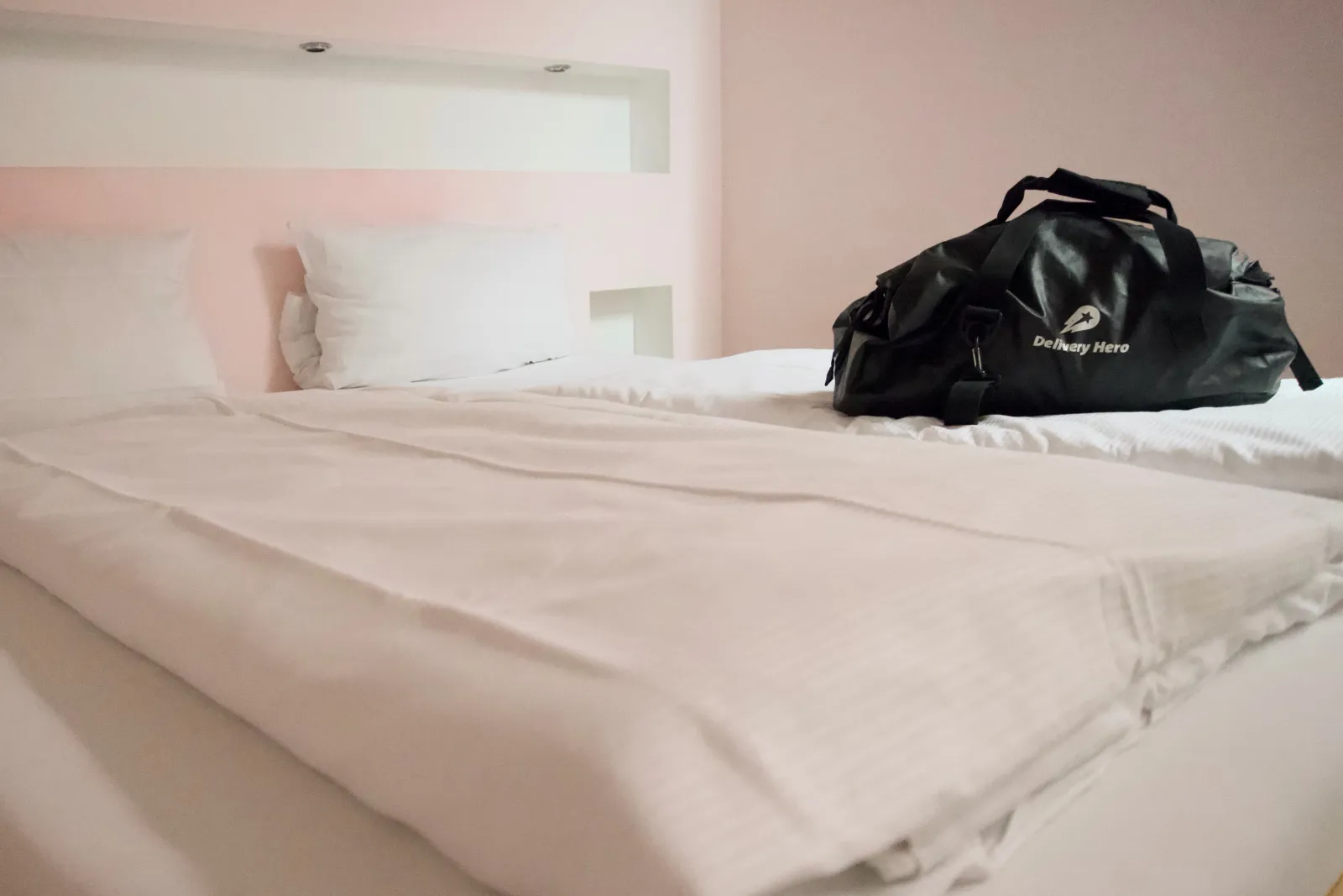 Black and white hotel room view of a freshly made bed with crisp white linen and two pillows, a black Delivery Hero duffel bag placed on the corner