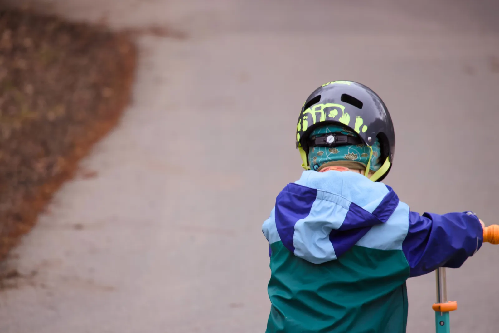 Small child from behind on a balance bike, wearing a dark helmet and blue rain jacket