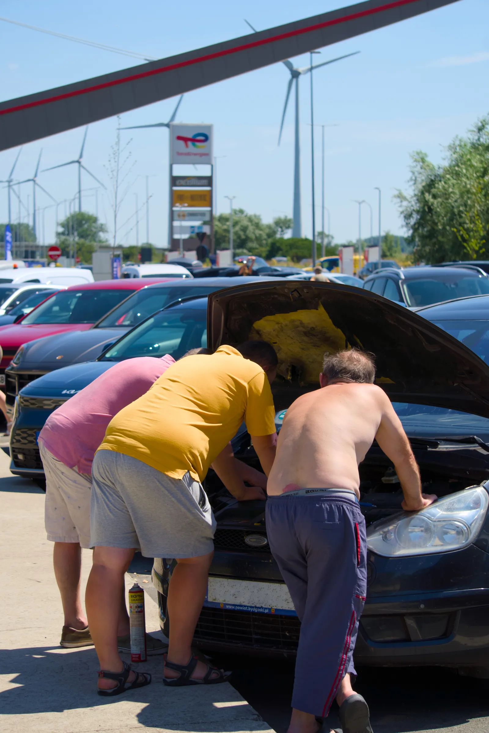 Three men under the open hood of a car at a service area, wind turbines behind