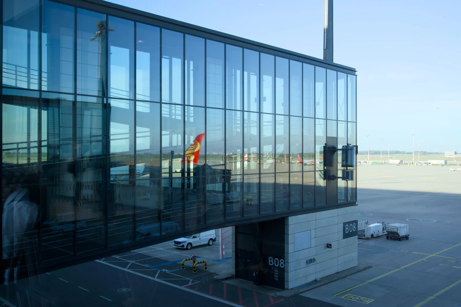 Boarding gate B08 seen through reflective glass, with a plane tail visible outside and a white van parked below