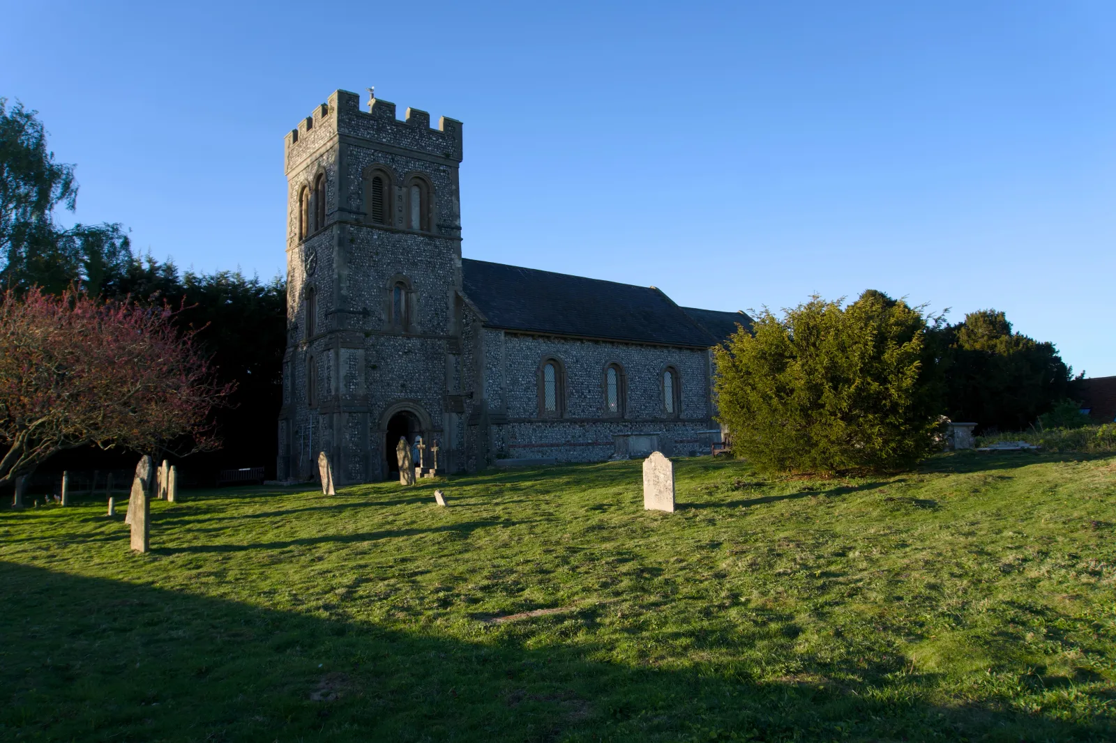 A flint stone village church with a square tower and slate roof, lit by low evening sun, gravestones scattered across the sloping lawn