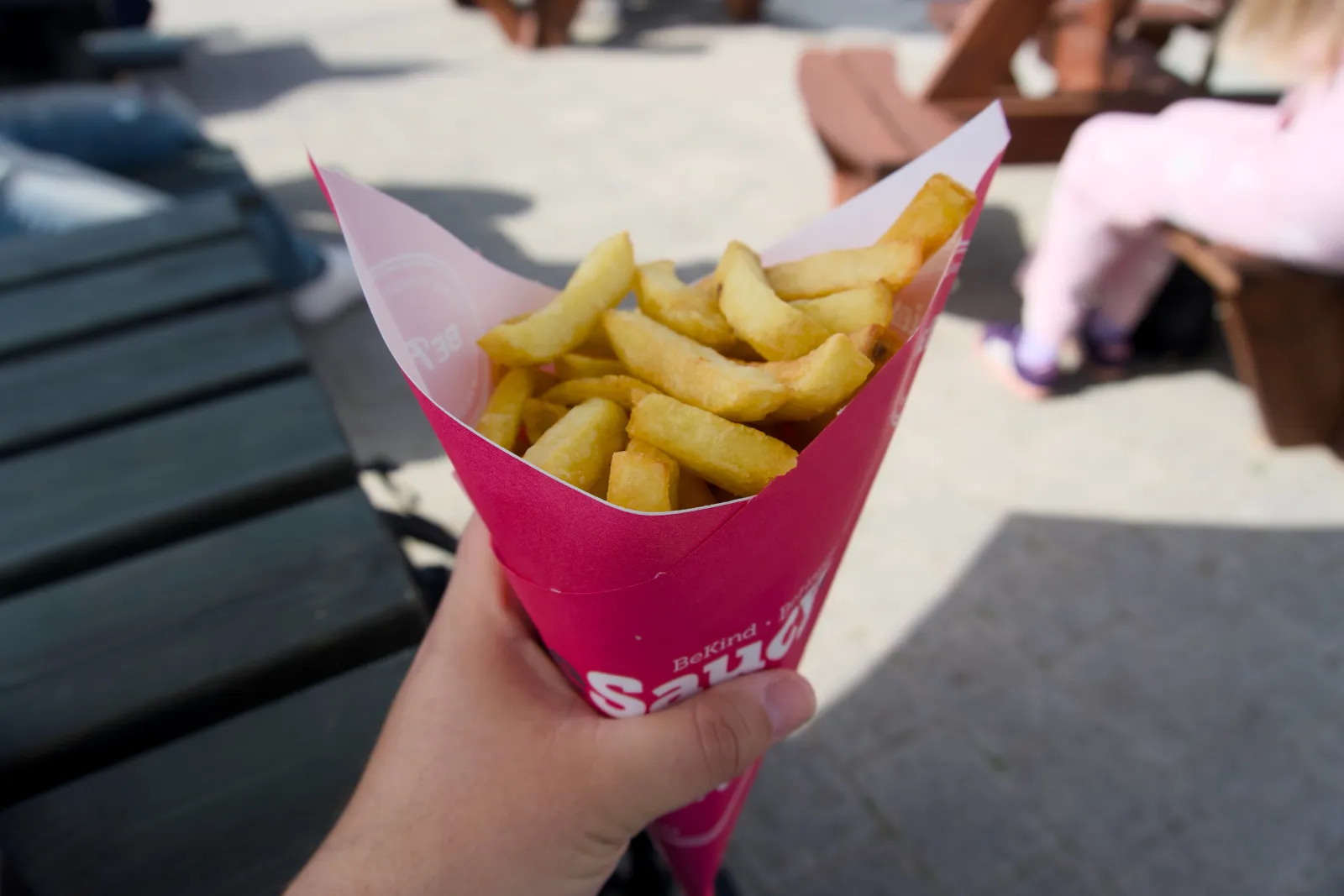 A hand holding a tall pink paper cone of thick-cut chips labelled 'Saucy', sunlit on the seafront with people sitting on benches in the background