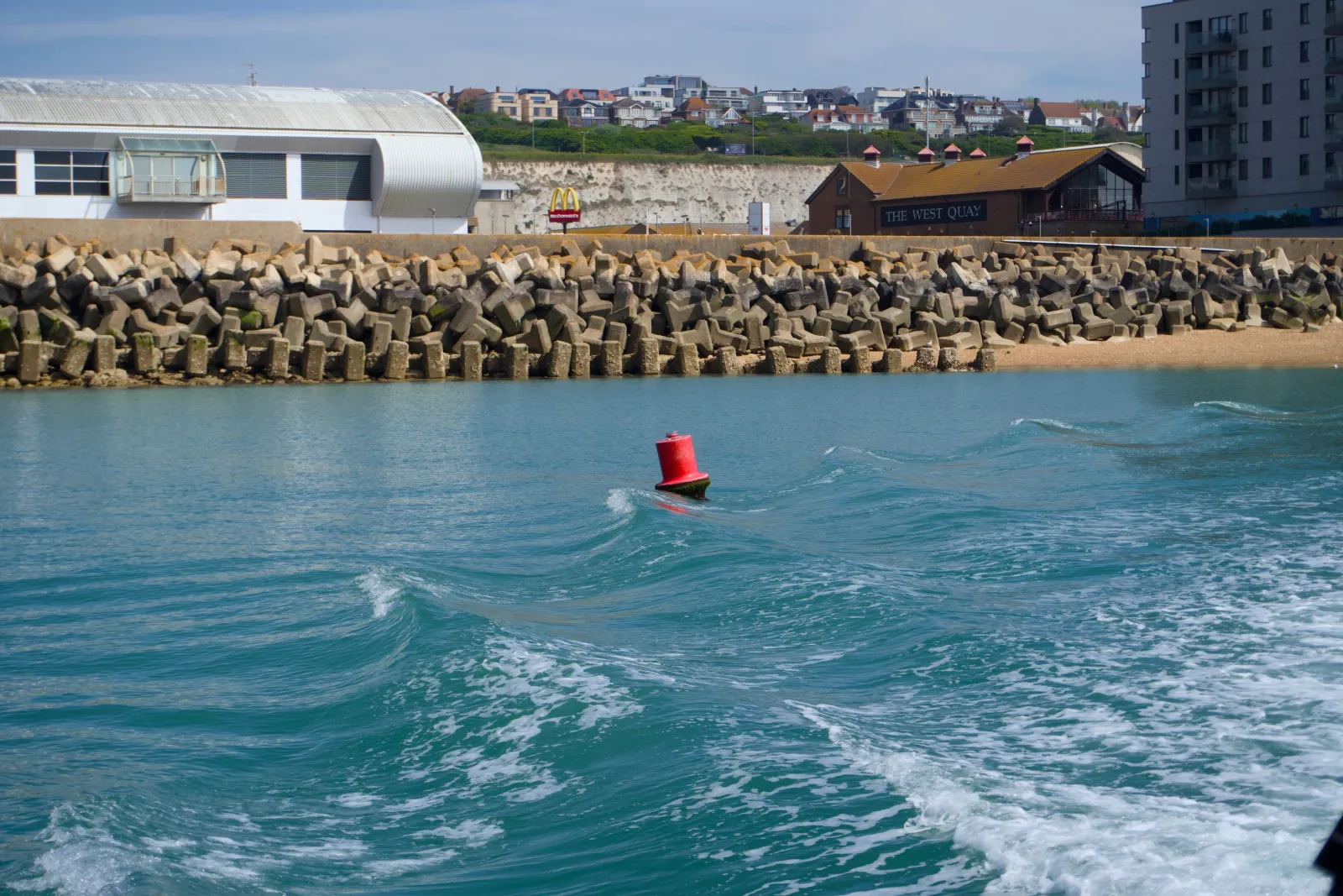 A red metal channel buoy floating in front of a stacked concrete sea wall, with chalk cliffs and the Brighton seafront visible behind