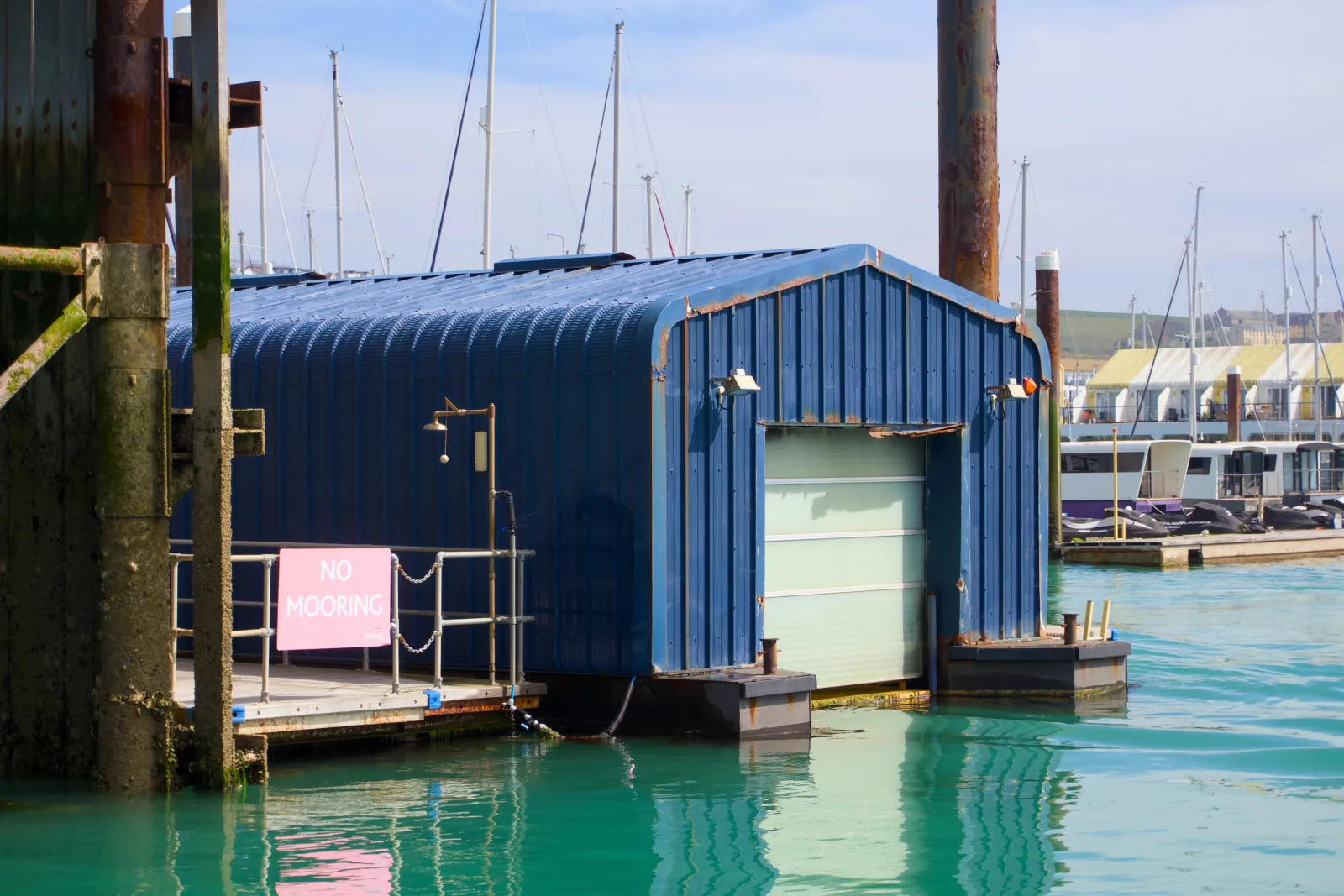 A blue corrugated metal boat house with closed roller door at Brighton Marina, a pink 'NO MOORING' sign on the railing, mast tops and the white cliffs visible in the background