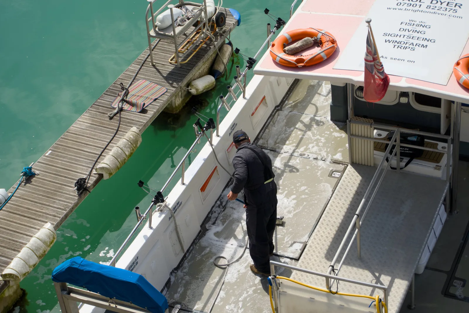 View from above of a man in dark workwear hosing down the deck of a fishing and sightseeing boat tied up at a pontoon, with mooring lines and ropes around the deck