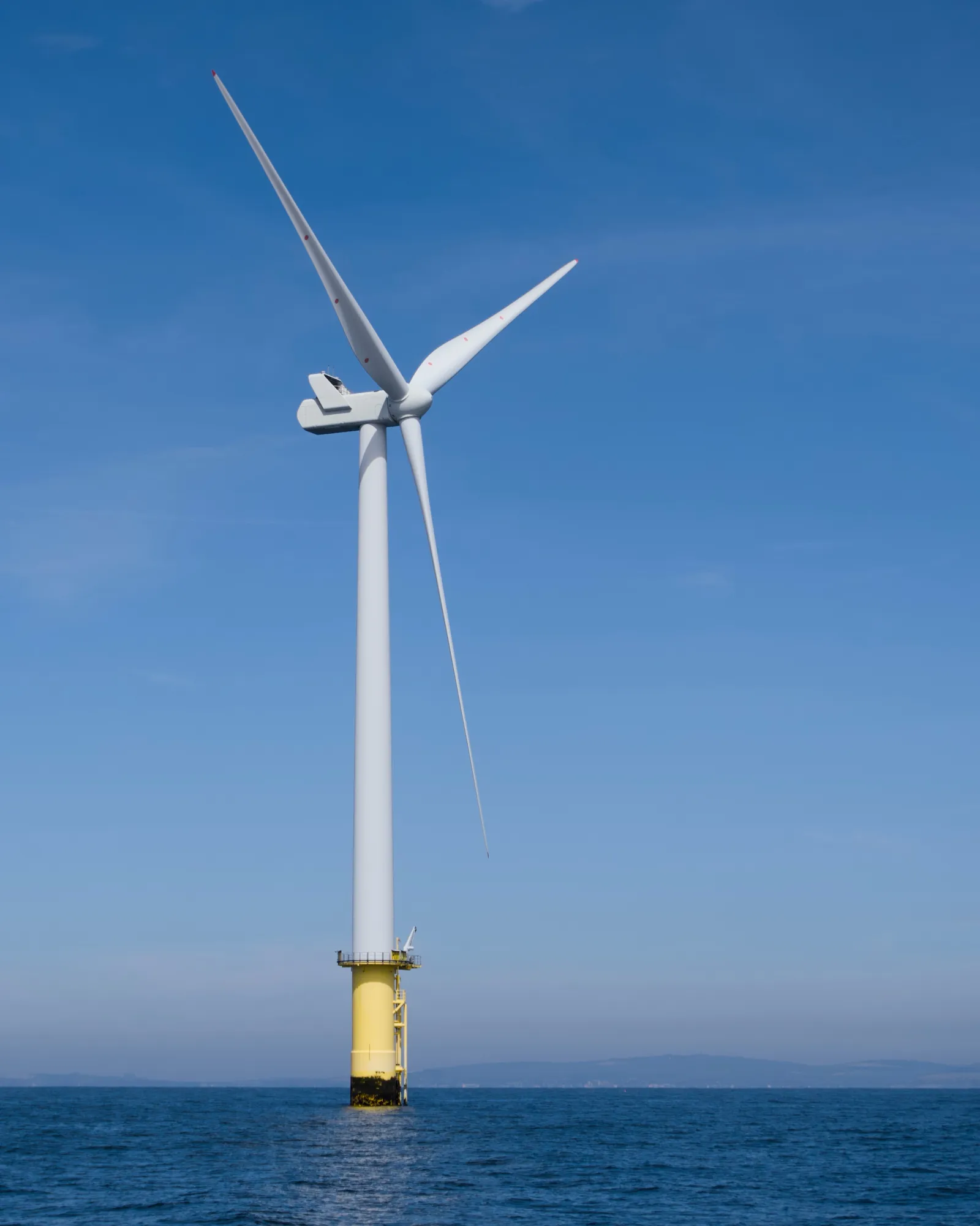 A full offshore wind turbine standing on a yellow transition piece in the sea, three white blades against a clear blue sky, the coast faintly visible on the horizon