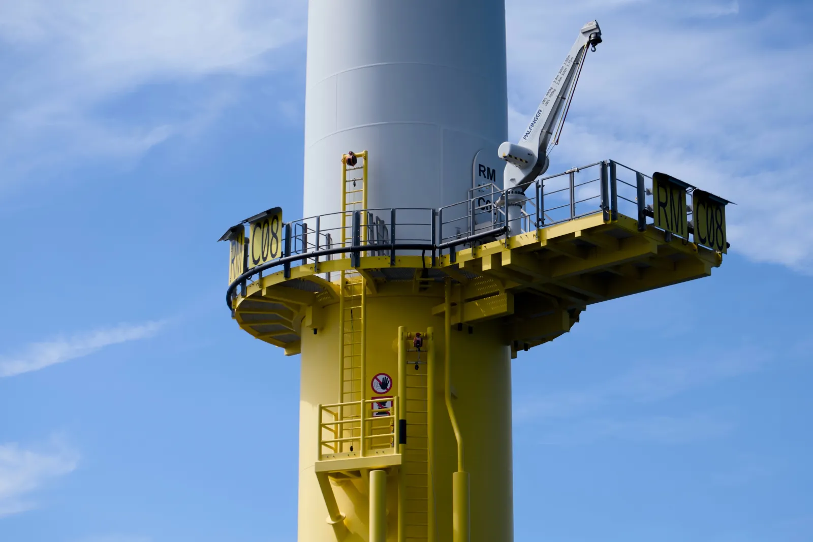 Close-up of an offshore wind turbine base with the yellow access platform, ladder and a service crane, marked RM C08, against a blue sky with thin clouds