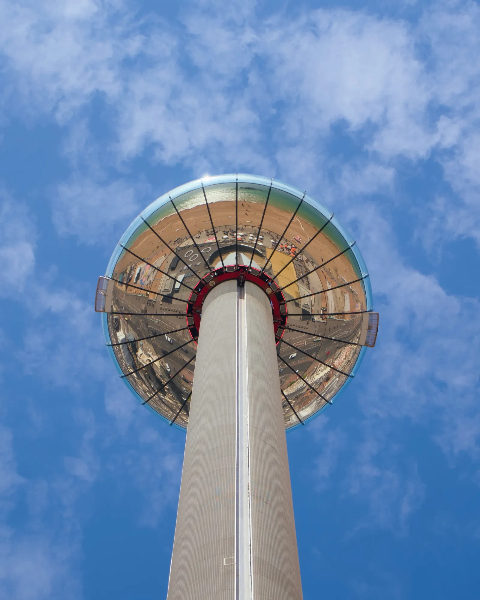 Looking straight up at the Brighton i360 observation tower, the glass viewing pod docked at the top reflecting the seafront, against a partly cloudy blue sky