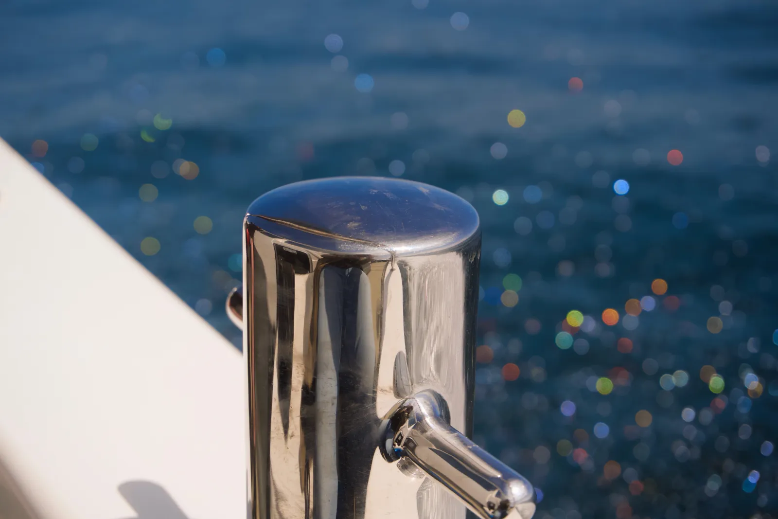 Close-up of a polished stainless steel mooring cleat on a white boat, with sparkling sun reflections on the dark blue sea behind it