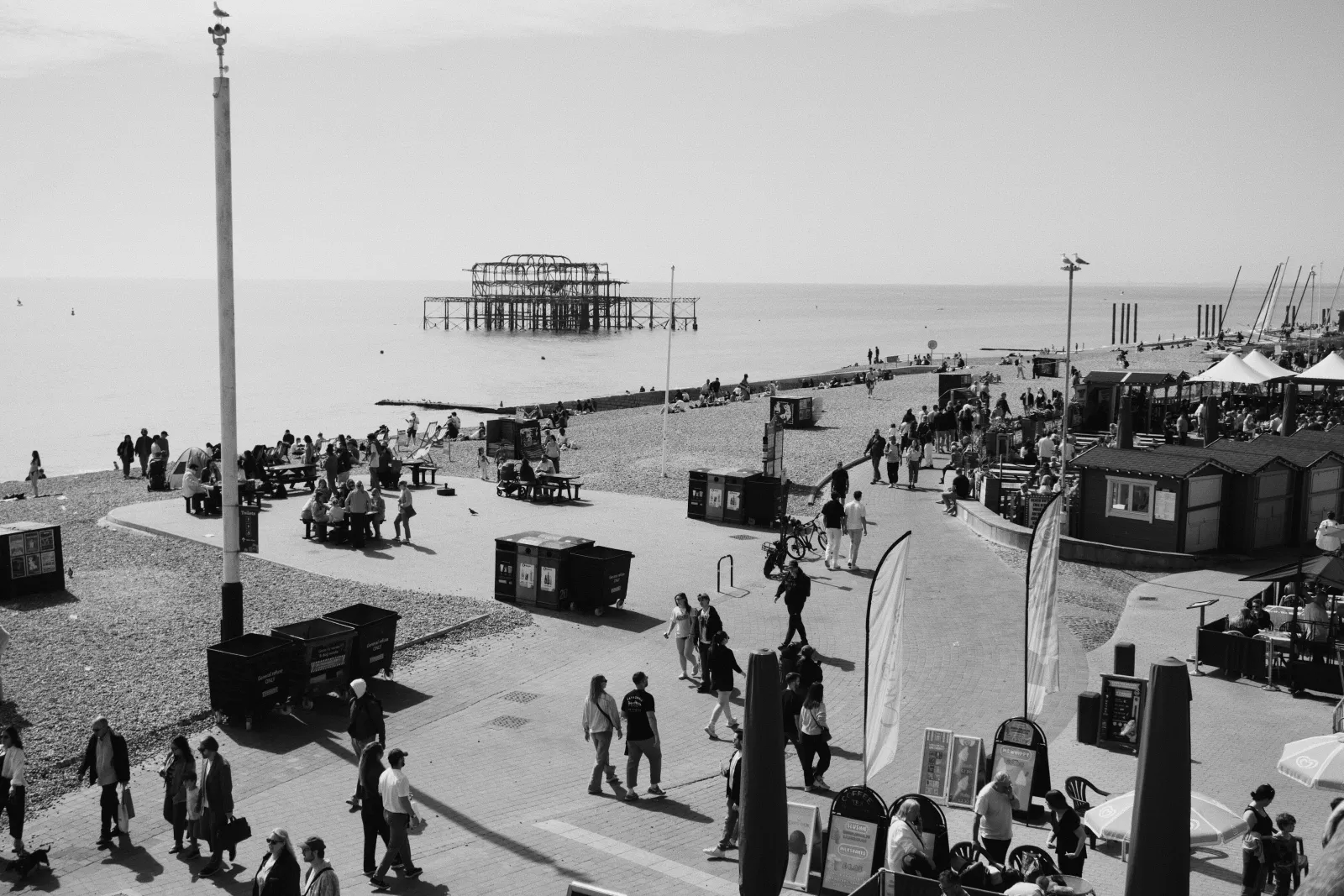 Black and white view of Brighton seafront promenade with crowds, food stalls, benches and the burnt-out skeleton of the West Pier sitting in the sea behind
