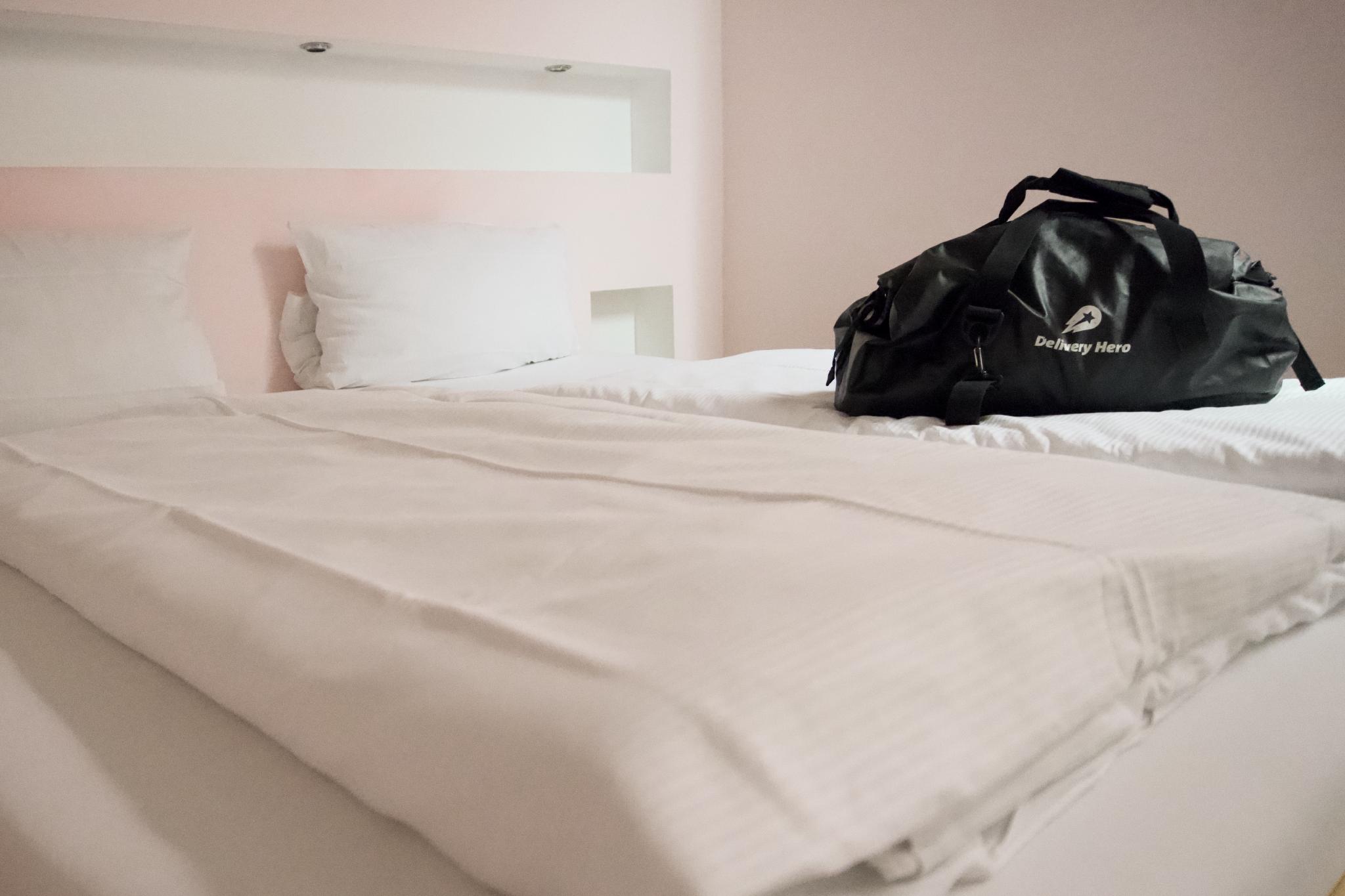 Black and white hotel room view of a freshly made bed with crisp white linen and two pillows, a black Delivery Hero duffel bag placed on the corner