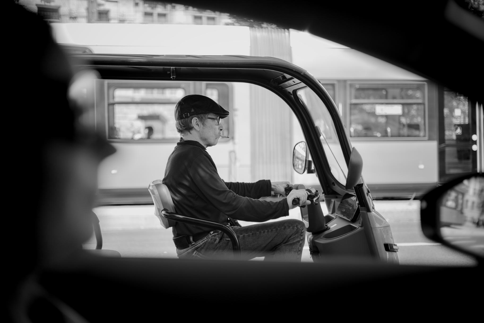 Black and white photo of a man with a flat cap driving an electric rickshaw