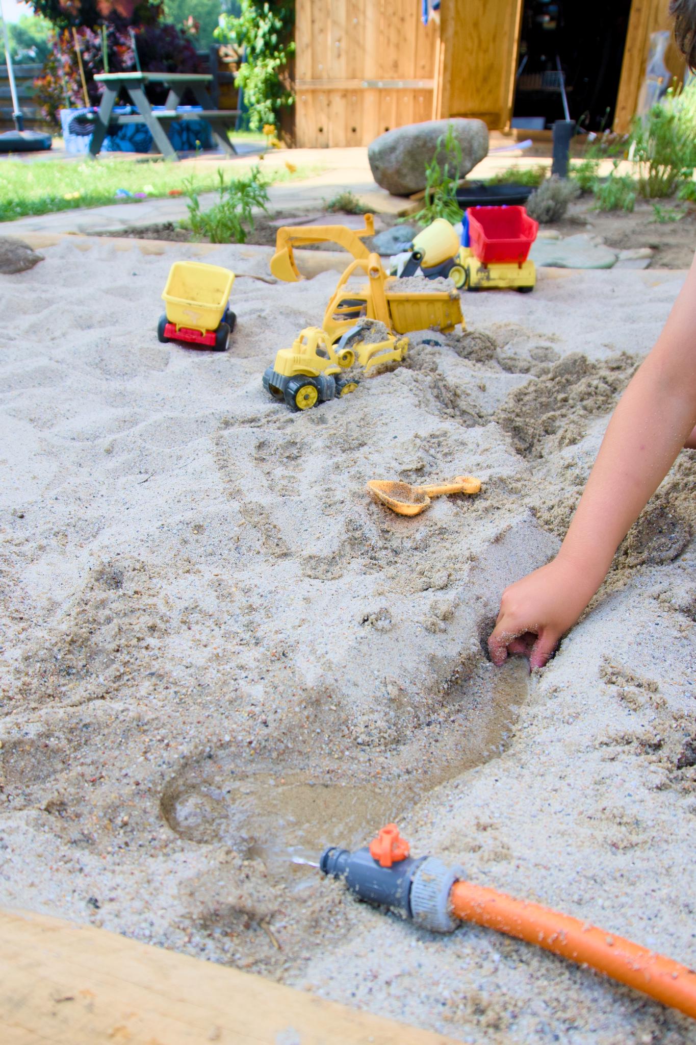 Child's hand digging in a sandpit with toy diggers and a water hose