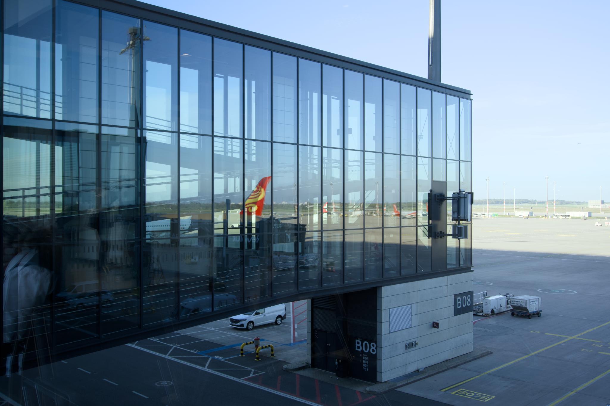 Boarding gate B08 seen through reflective glass, with a plane tail visible outside and a white van parked below