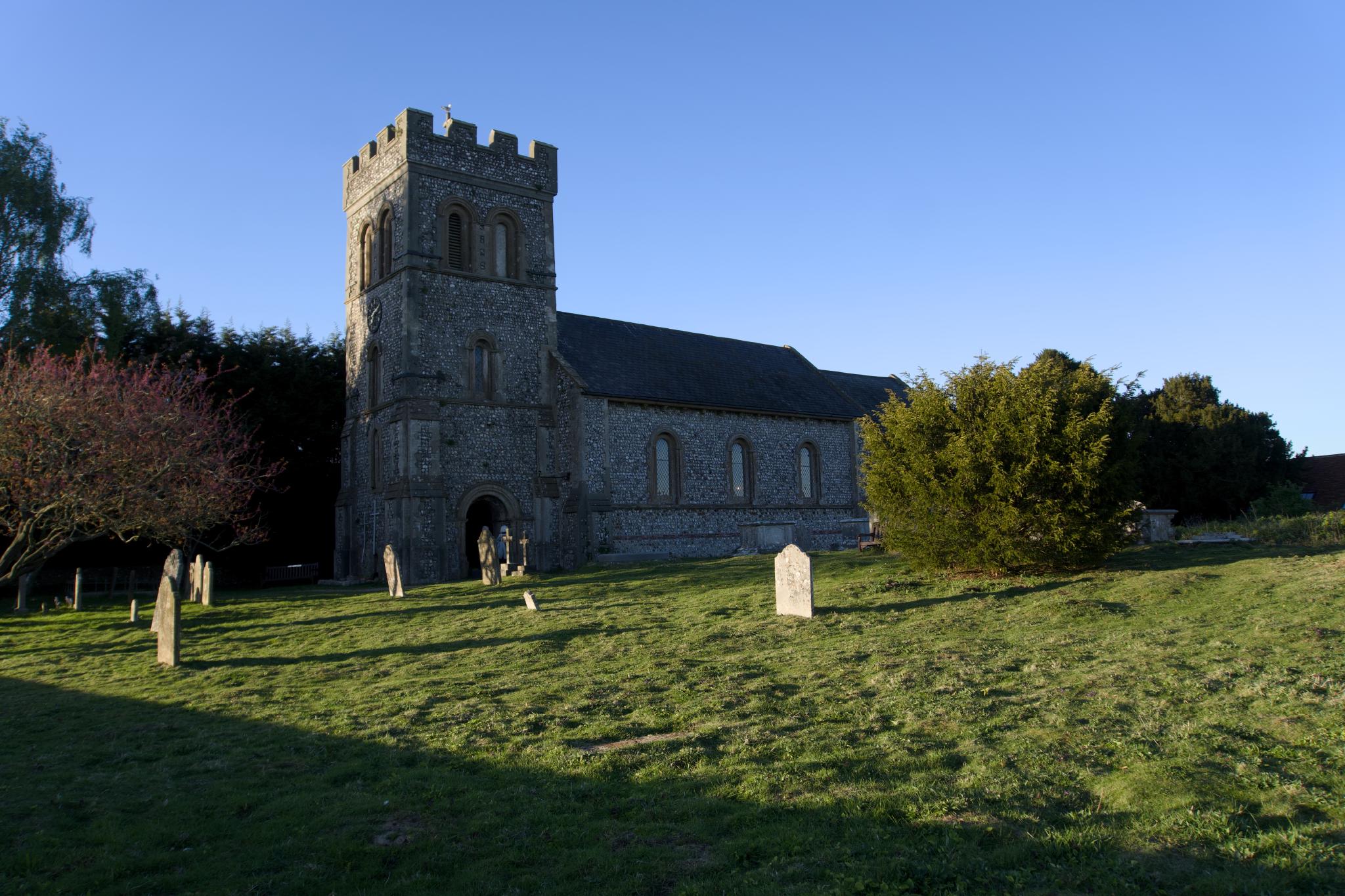 A flint stone village church with a square tower and slate roof, lit by low evening sun, gravestones scattered across the sloping lawn