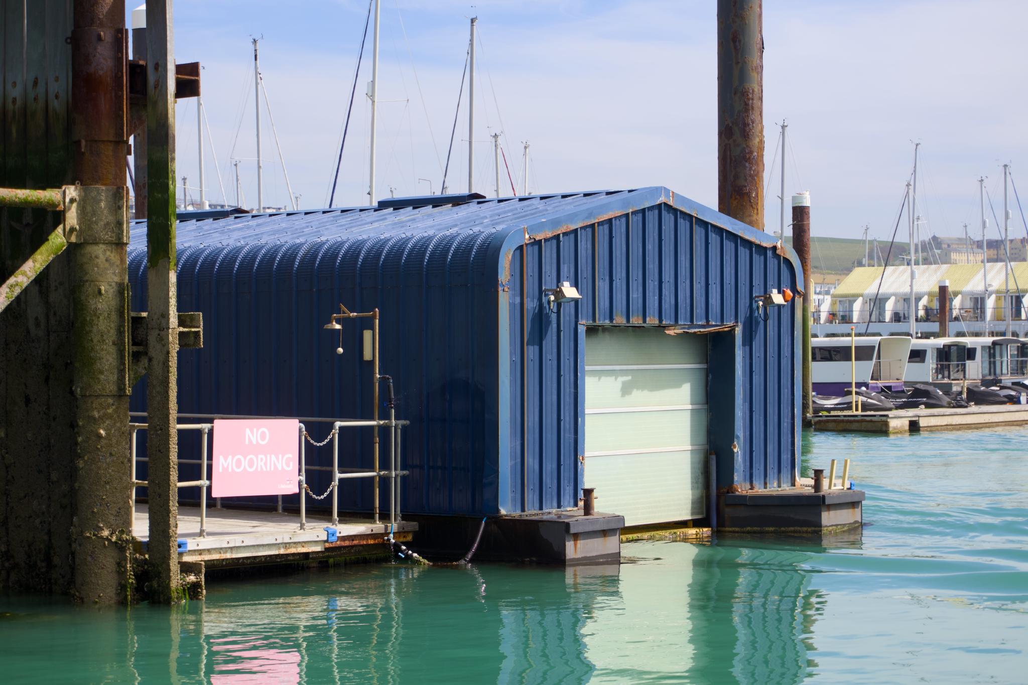 A blue corrugated metal boat house with closed roller door at Brighton Marina, a pink 'NO MOORING' sign on the railing, mast tops and the white cliffs visible in the background