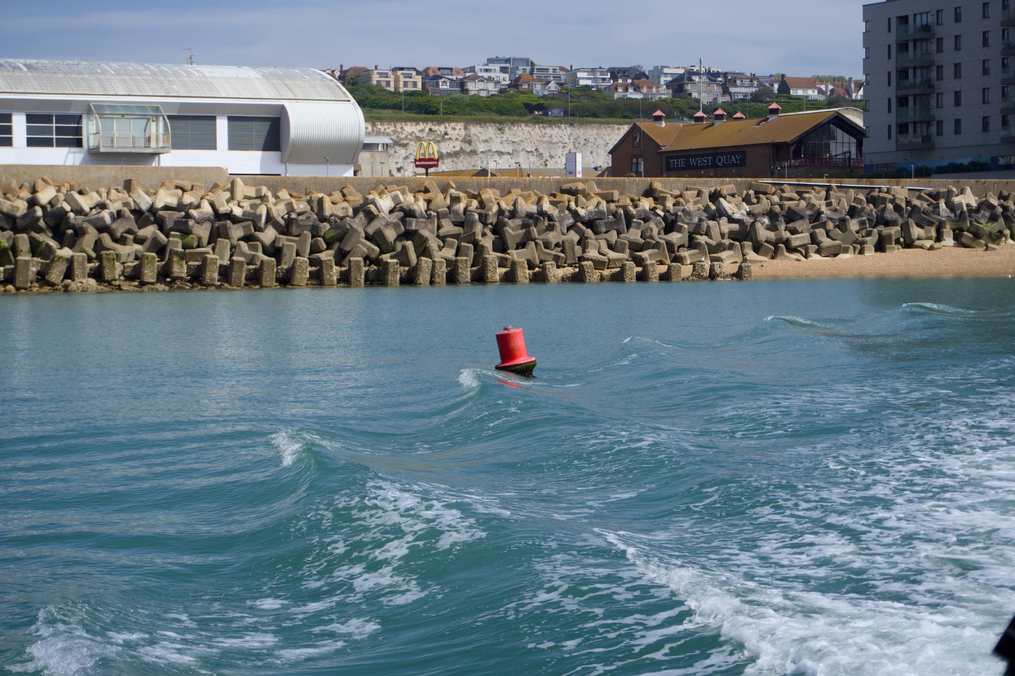A red metal channel buoy floating in front of a stacked concrete sea wall, with chalk cliffs and the Brighton seafront visible behind