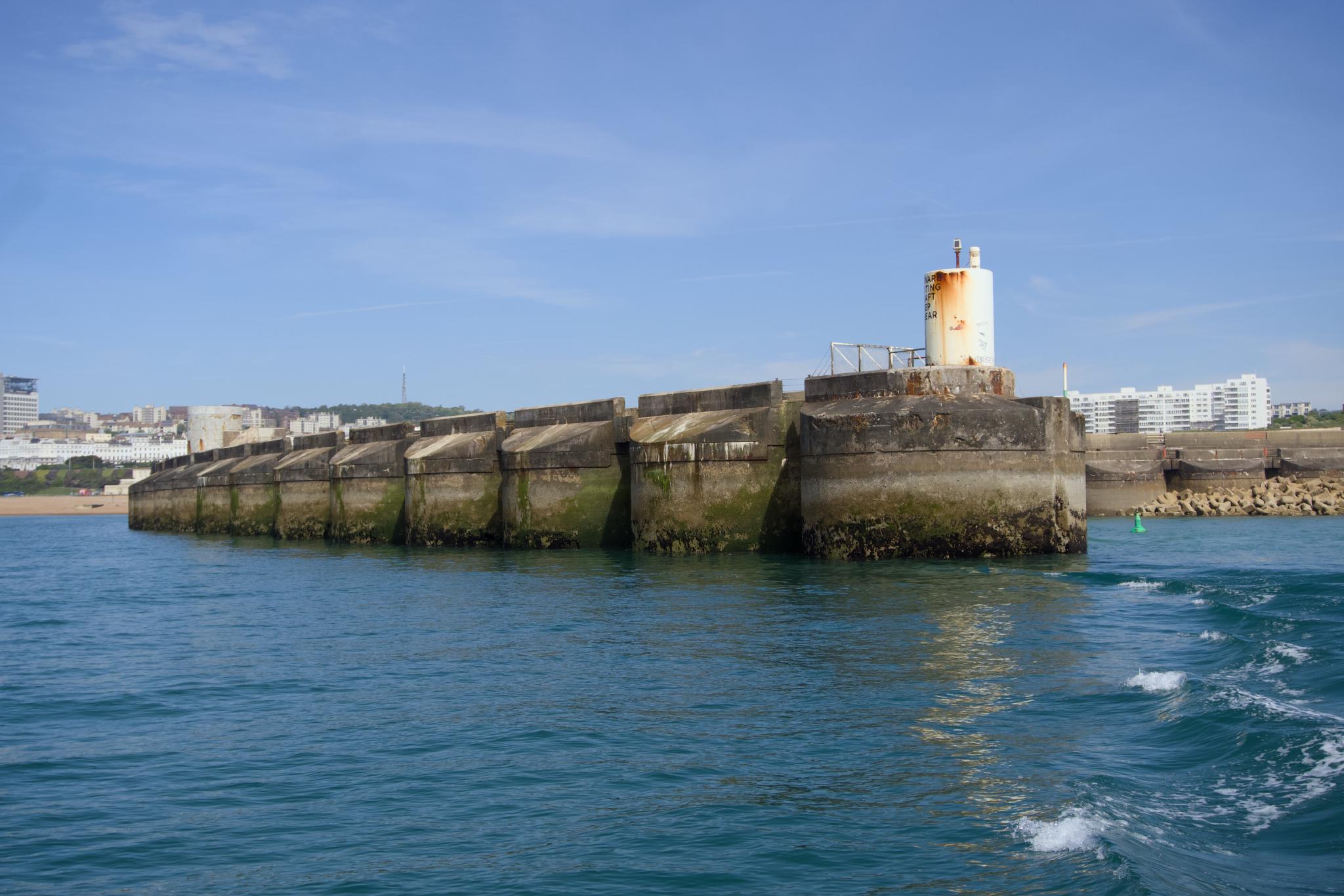 A long concrete sea wall with a small white and rust-stained light tower at the end, against a clear blue sky and turquoise sea