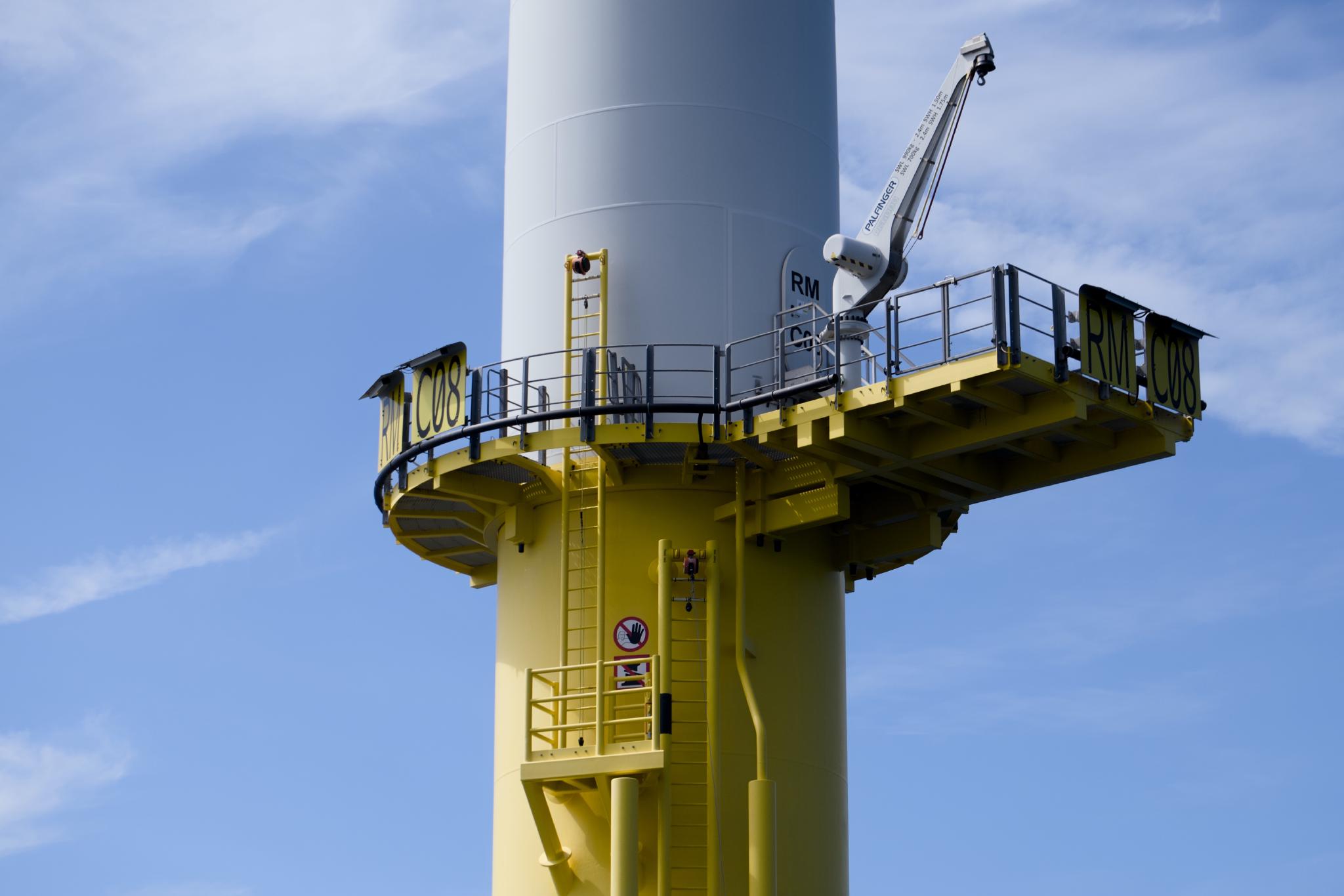 Close-up of an offshore wind turbine base with the yellow access platform, ladder and a service crane, marked RM C08, against a blue sky with thin clouds