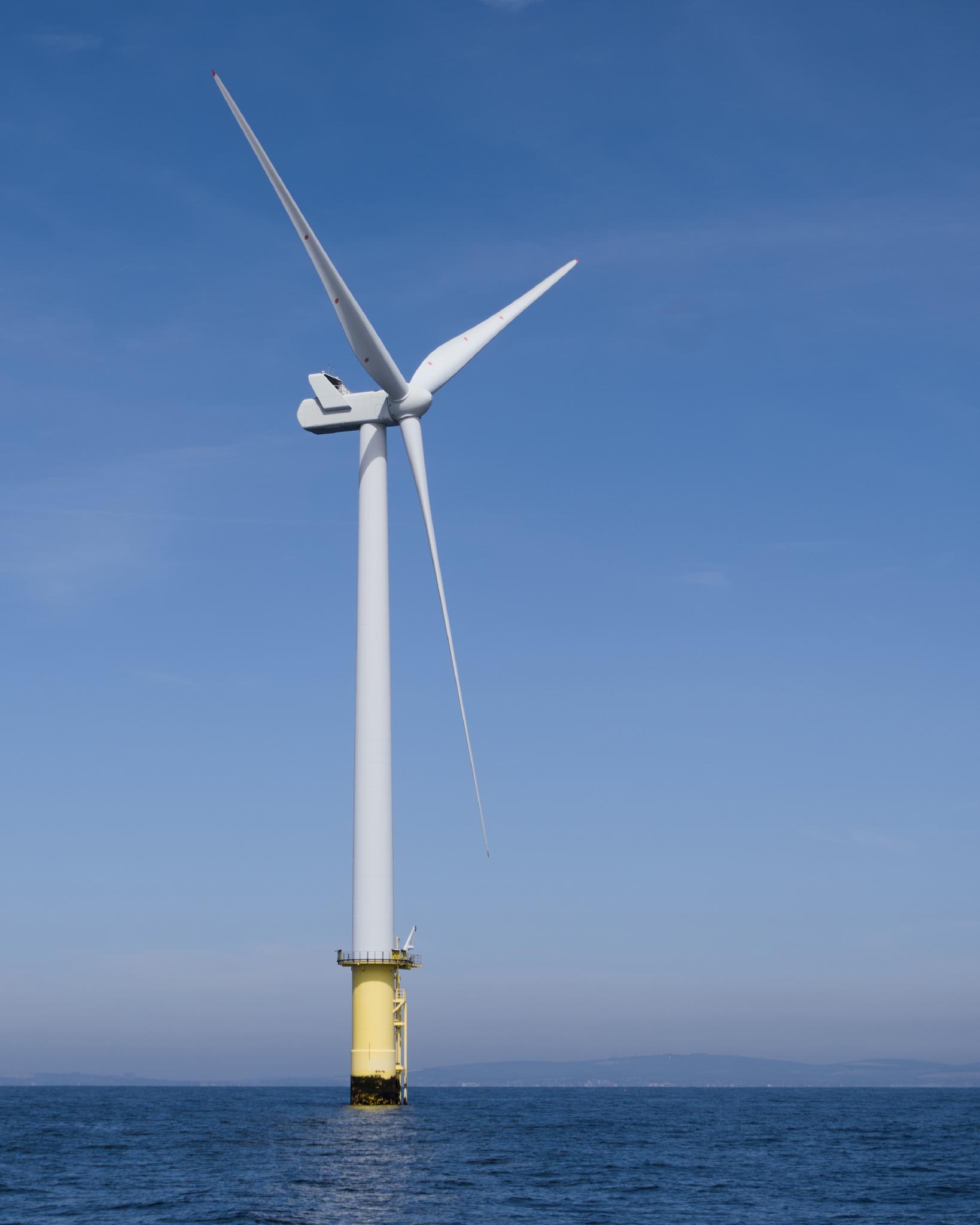 A full offshore wind turbine standing on a yellow transition piece in the sea, three white blades against a clear blue sky, the coast faintly visible on the horizon