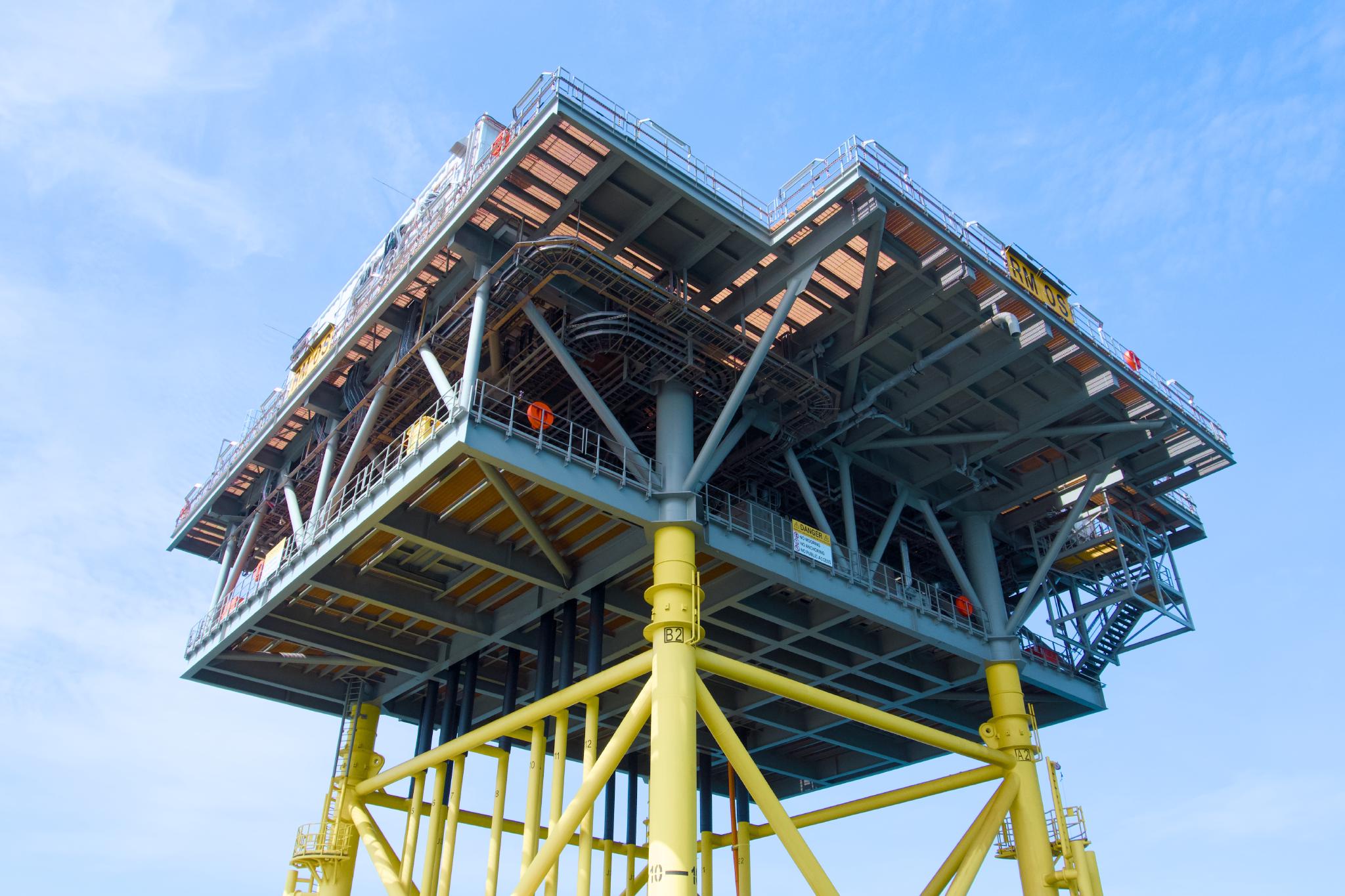 An offshore substation platform from below, blue steel deck supported by a yellow tubular jacket structure rising out of the sea