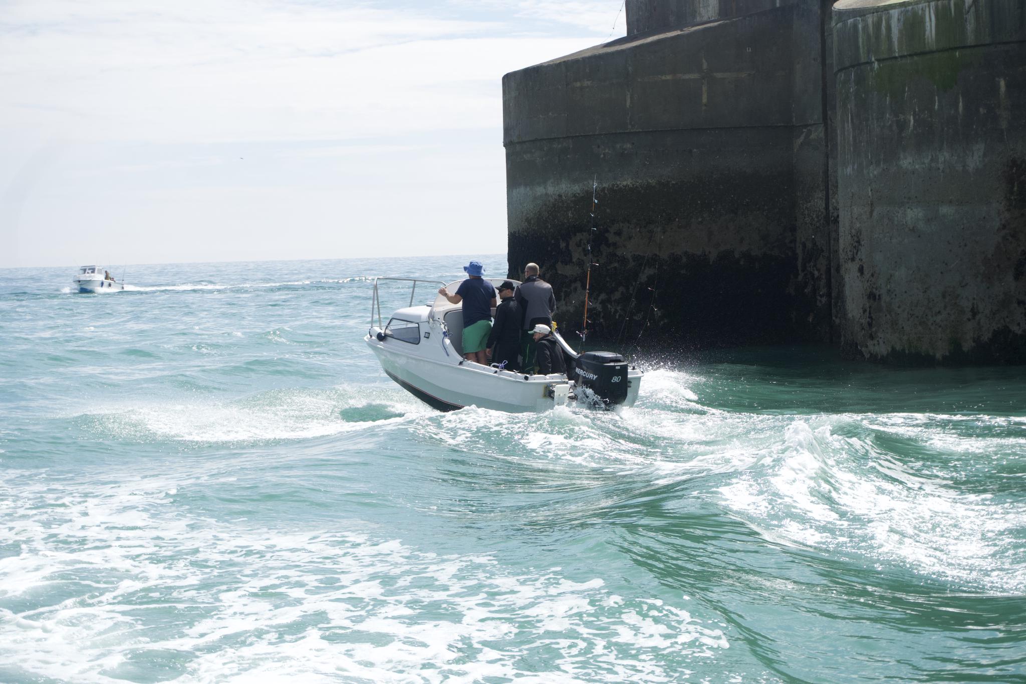 A small white fishing boat with three people on board returning to harbour past a tall dark sea wall, another boat in the distance