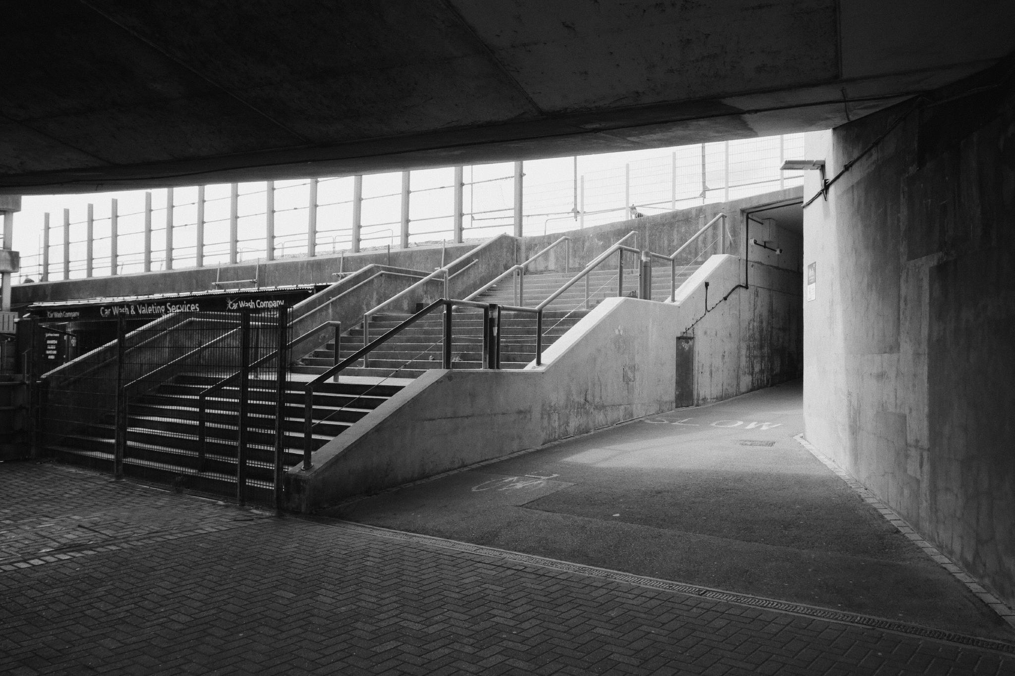 Black and white view of a deserted concrete underpass at a station with two flights of stairs leading up into bright daylight