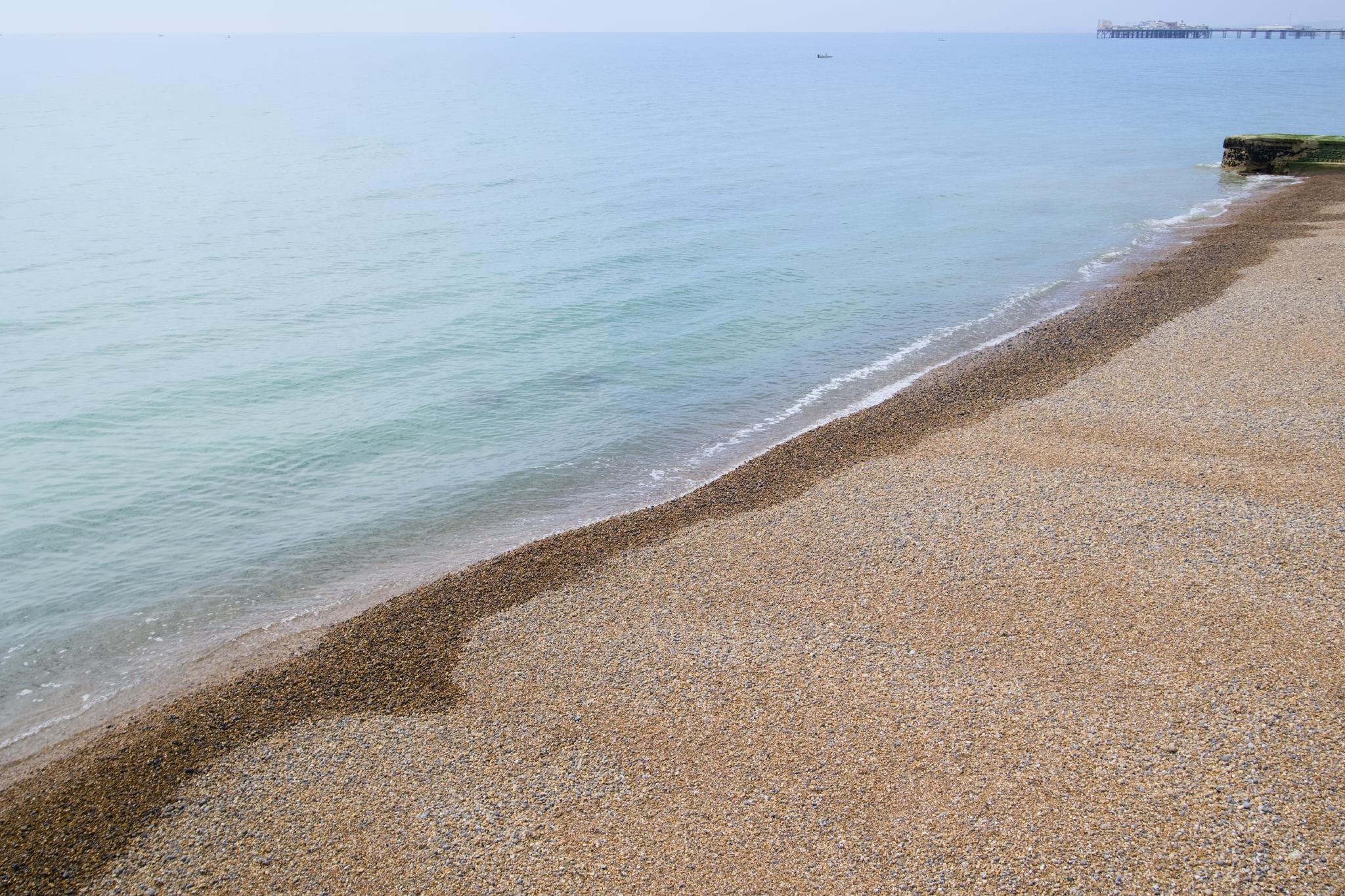 Brighton pebble beach in soft afternoon light, calm sea meeting the shoreline at a curved tideline, Brighton Palace Pier far in the distance