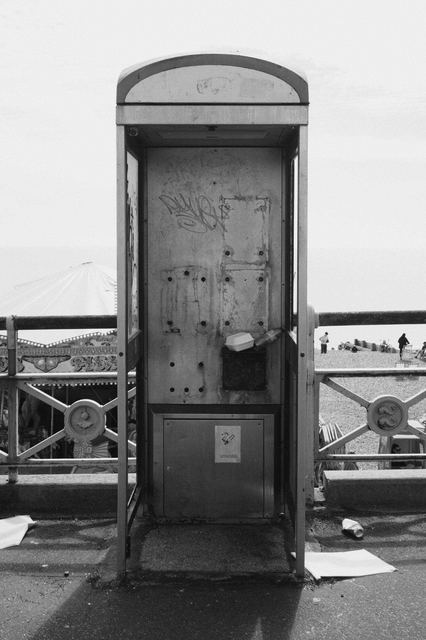 Black and white photograph of a battered, vandalised public phone booth on Brighton seafront, the receiver gone and graffiti scratched into the metal panels, beach and crowds visible behind