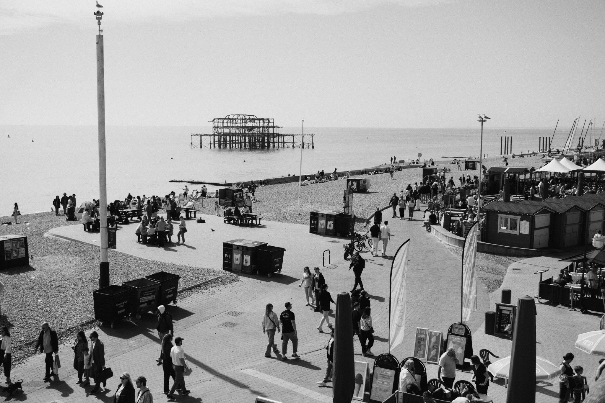 Black and white view of Brighton seafront promenade with crowds, food stalls, benches and the burnt-out skeleton of the West Pier sitting in the sea behind