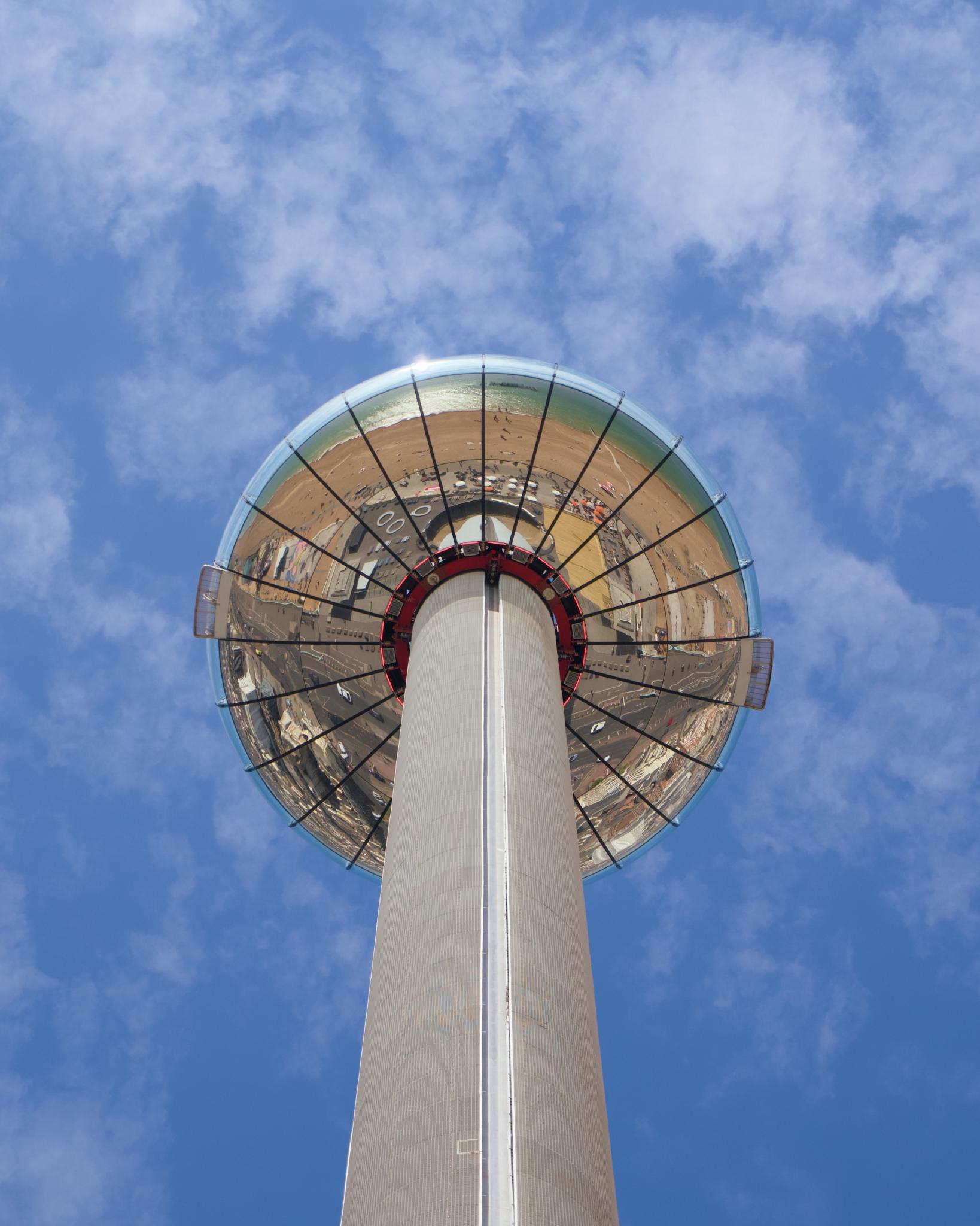 Looking straight up at the Brighton i360 observation tower, the glass viewing pod docked at the top reflecting the seafront, against a partly cloudy blue sky