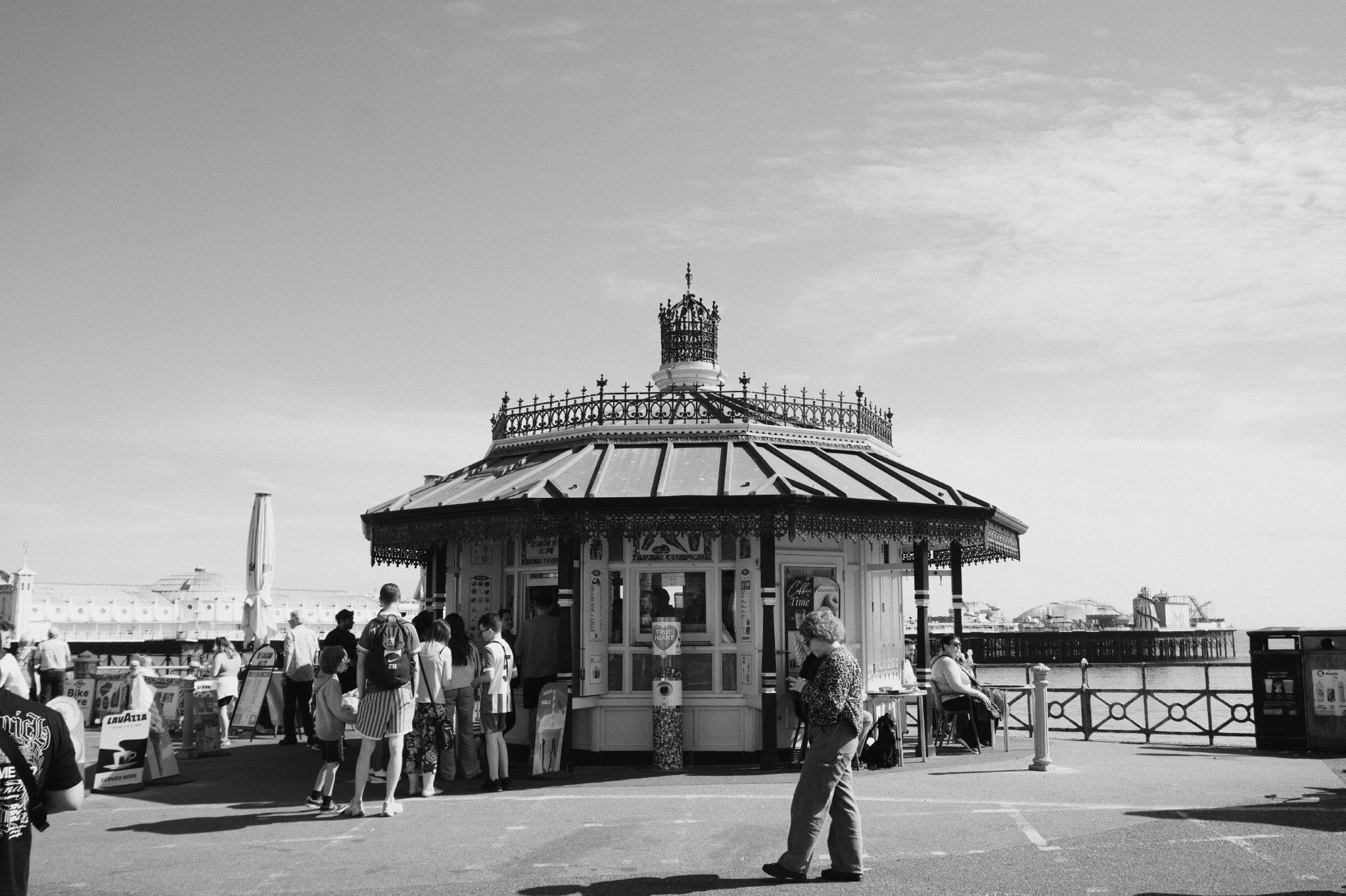 Black and white photograph of an ornate Victorian seafront kiosk on Brighton Pier with a domed crown roof, people queuing at the windows